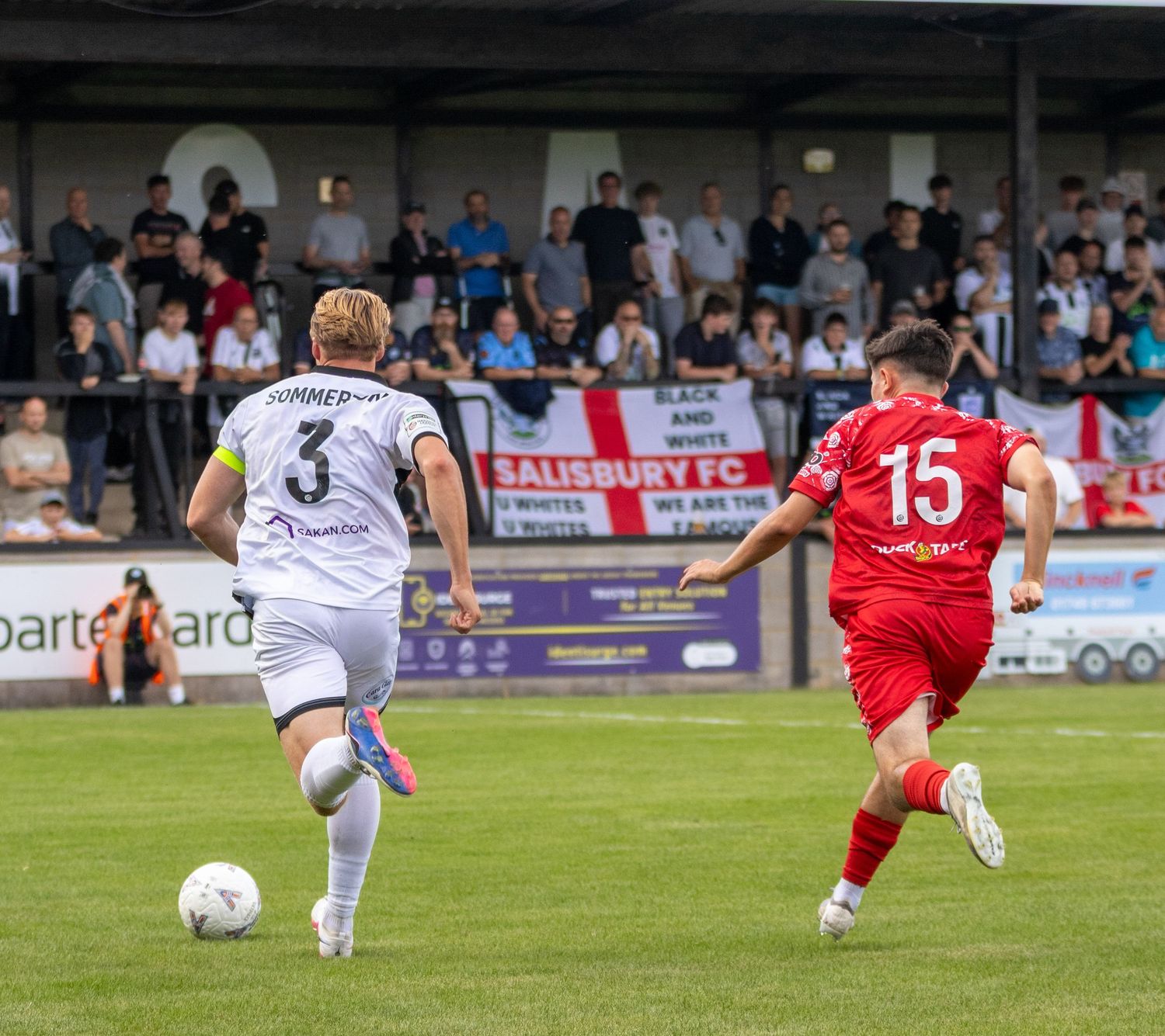 Two soccer players compete on the pitch at Salisbury FC stadium with spectators watching from the stands.