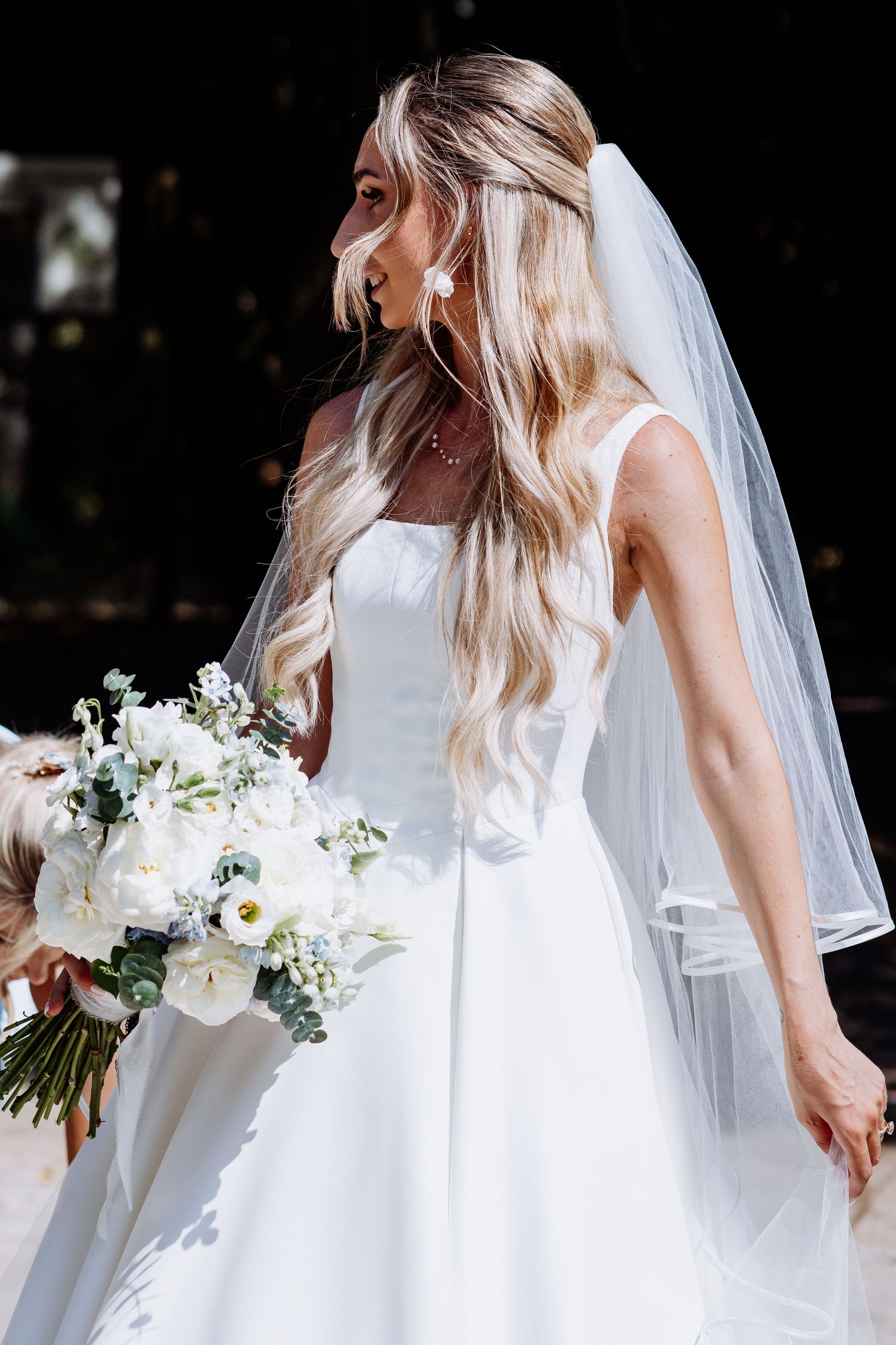 Elegant bride with long blonde hair holds a white floral bouquet wearing a sleek gown and veil.