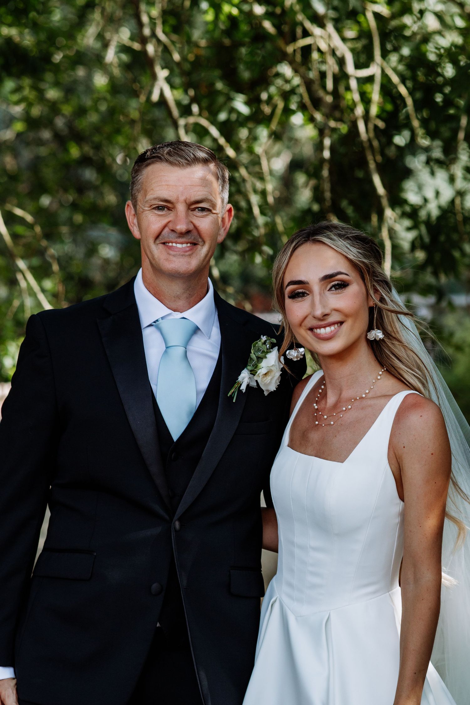 Smiling man in black tuxedo with light blue tie poses with bride in white dress at garden wedding.