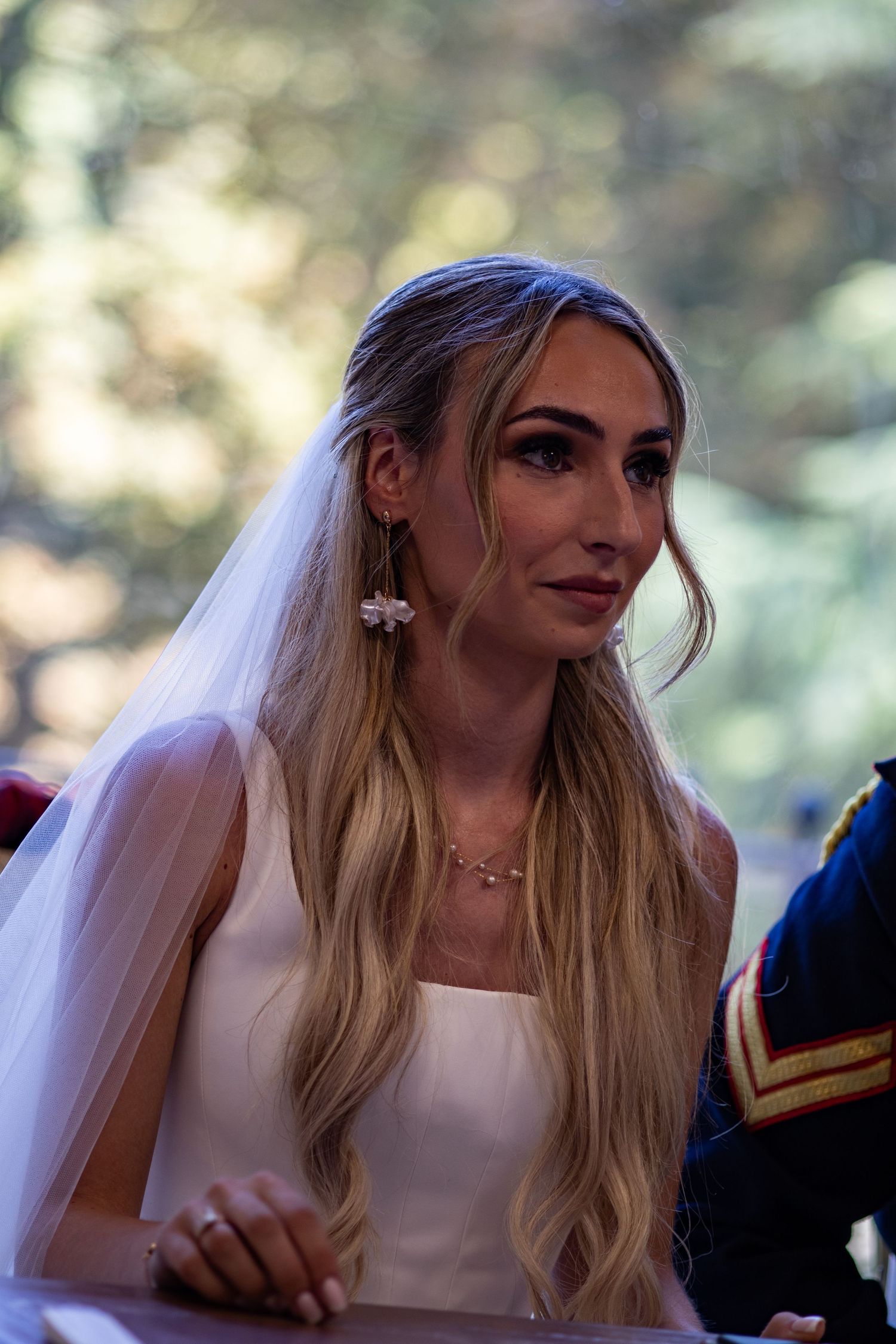 Beautiful blonde bride in white gown with veil and floral earrings smiling at wedding ceremony.