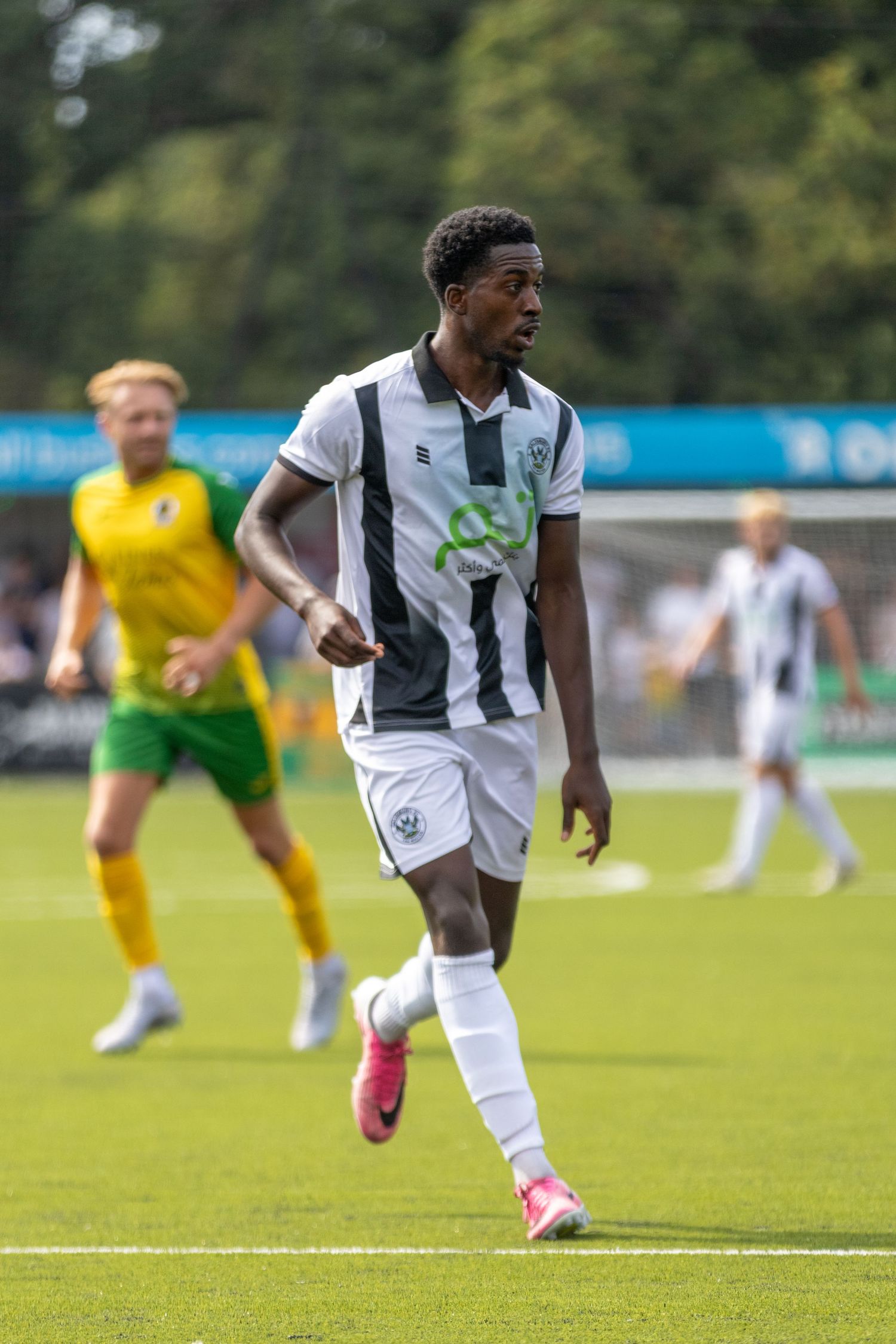 A footballer in black and white striped kit jogs across a green pitch during a sunny outdoor soccer match.