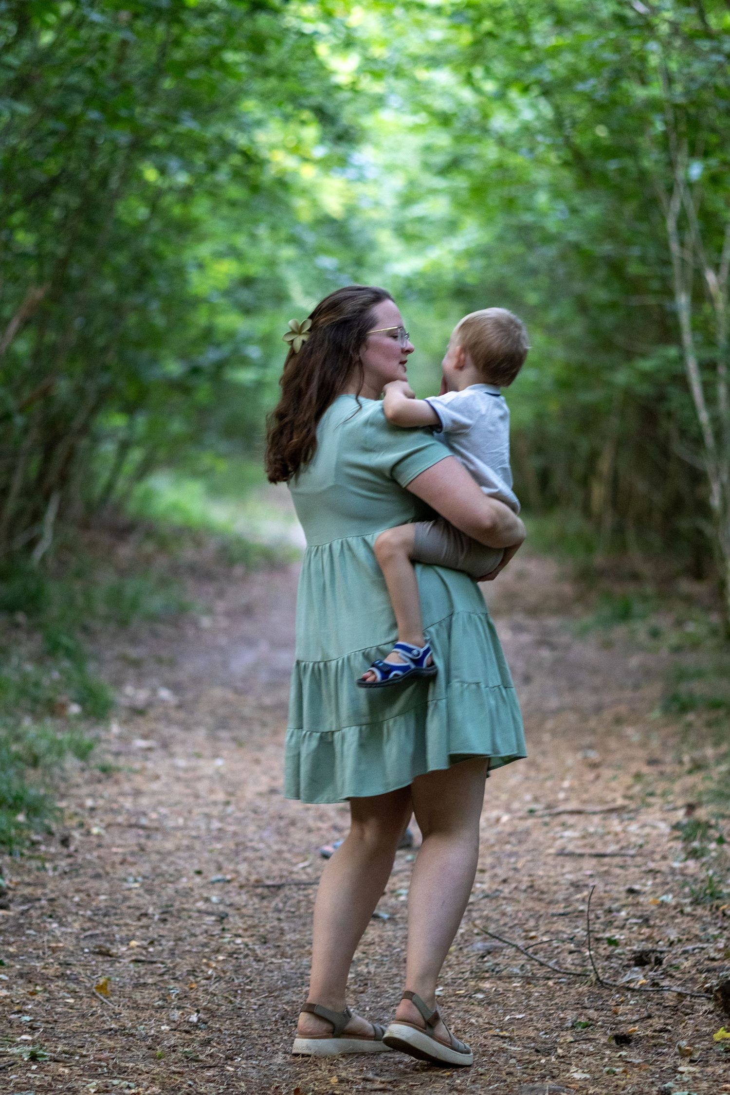 Mother in sage dress holding toddler while standing on a lush woodland trail.