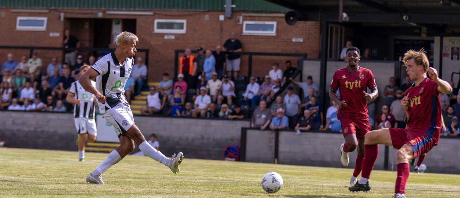 A footballer in black-white kit shoots at goal during a non-league match watched by a packed crowd.