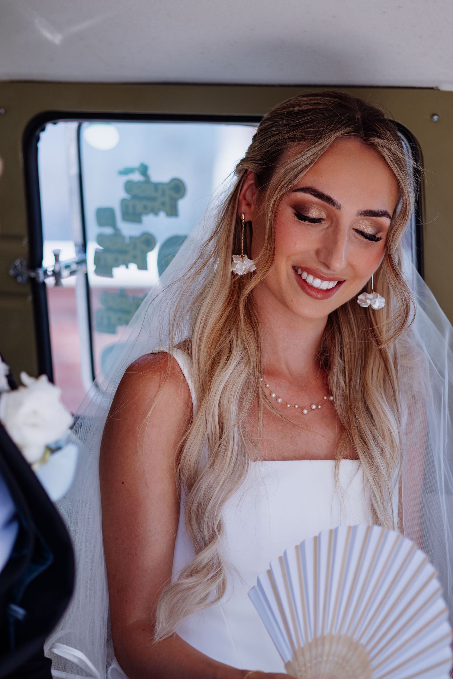 Radiant bride in white dress and veil holding a fan, wearing floral earrings inside a vintage vehicle.