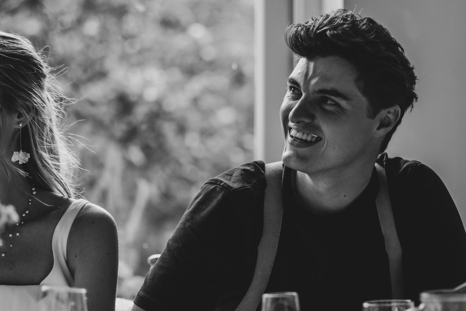 Black and white candid of laughing young man in black shirt seated beside blonde woman at wedding reception.