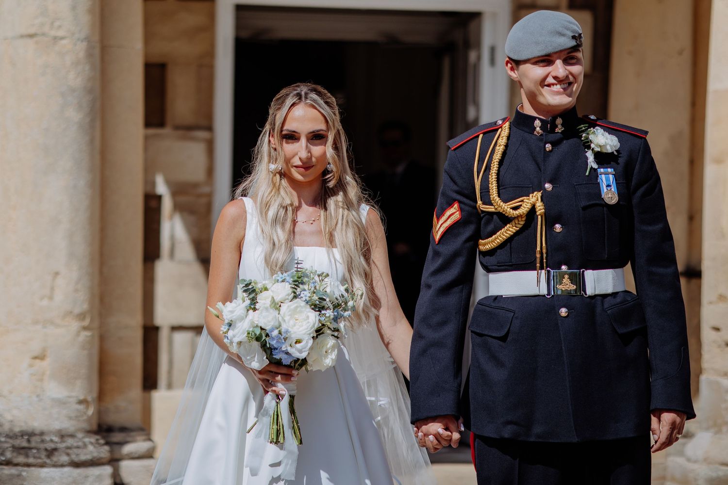 Newlywed couple walks hand in hand, bride in white dress with bouquet, groom in RAF dress uniform with beret.