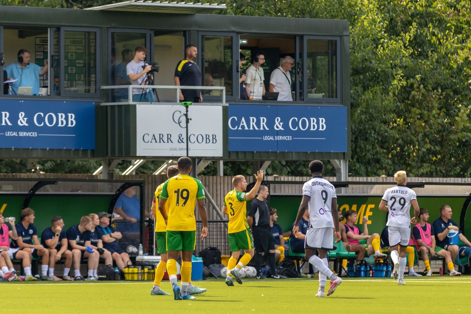 Football players in yellow-green and white kits compete near the Carr & Cobb sponsored stand.