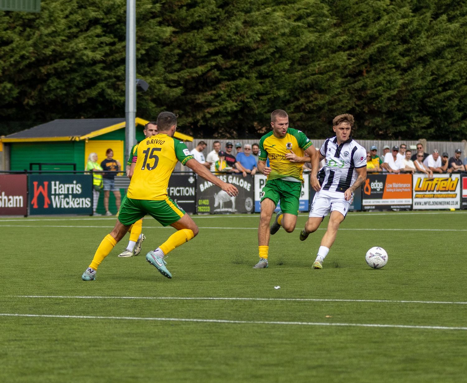 Players in yellow-green and white kits compete for the ball during a sunny non-league football match.
