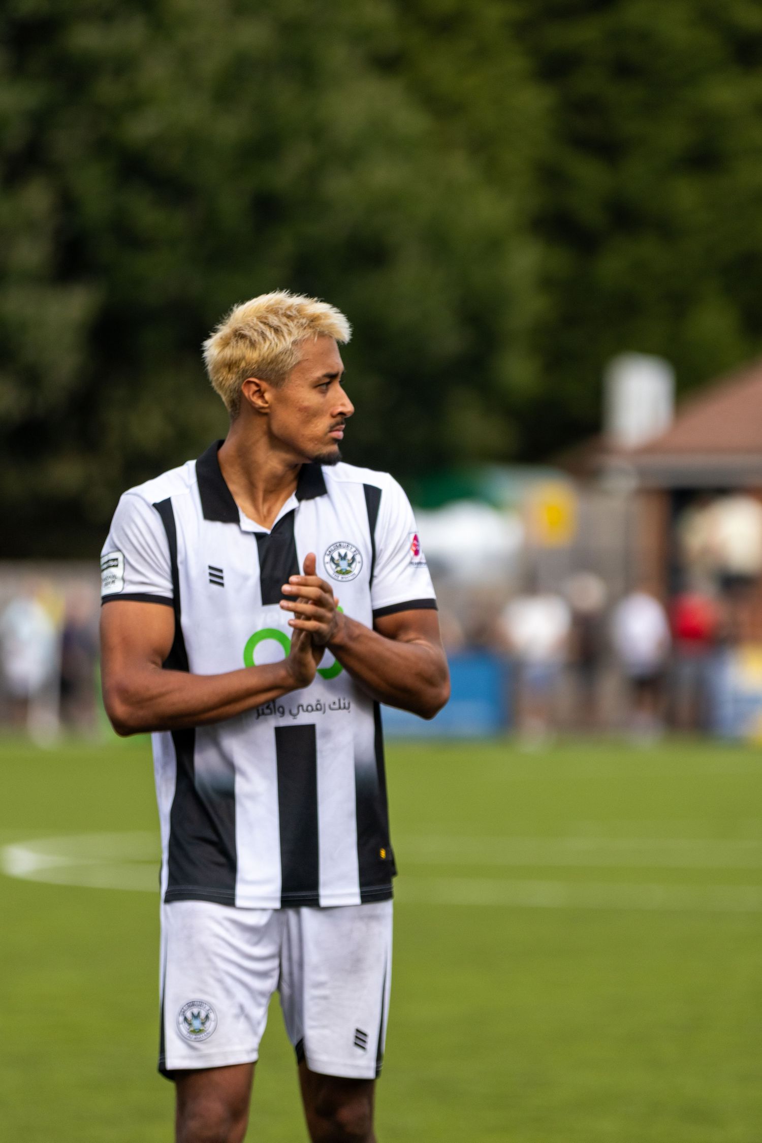 Blonde male footballer in black and white striped kit stands clapping on a grass pitch outdoors.