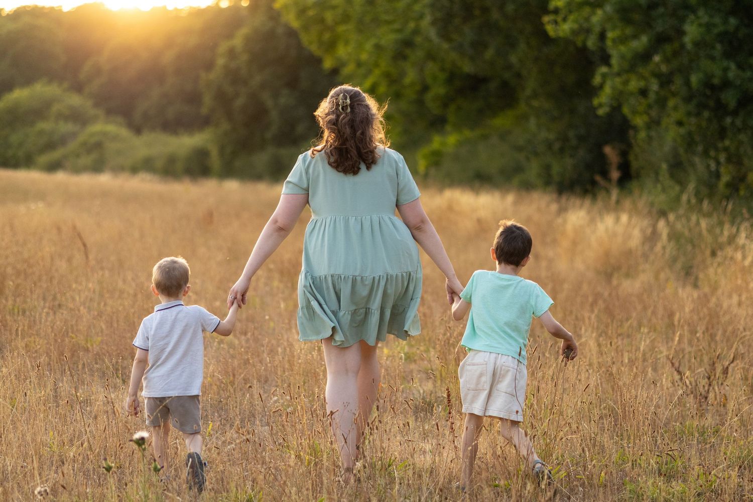 Family of mother and two young boys walking together through golden field at sunset.