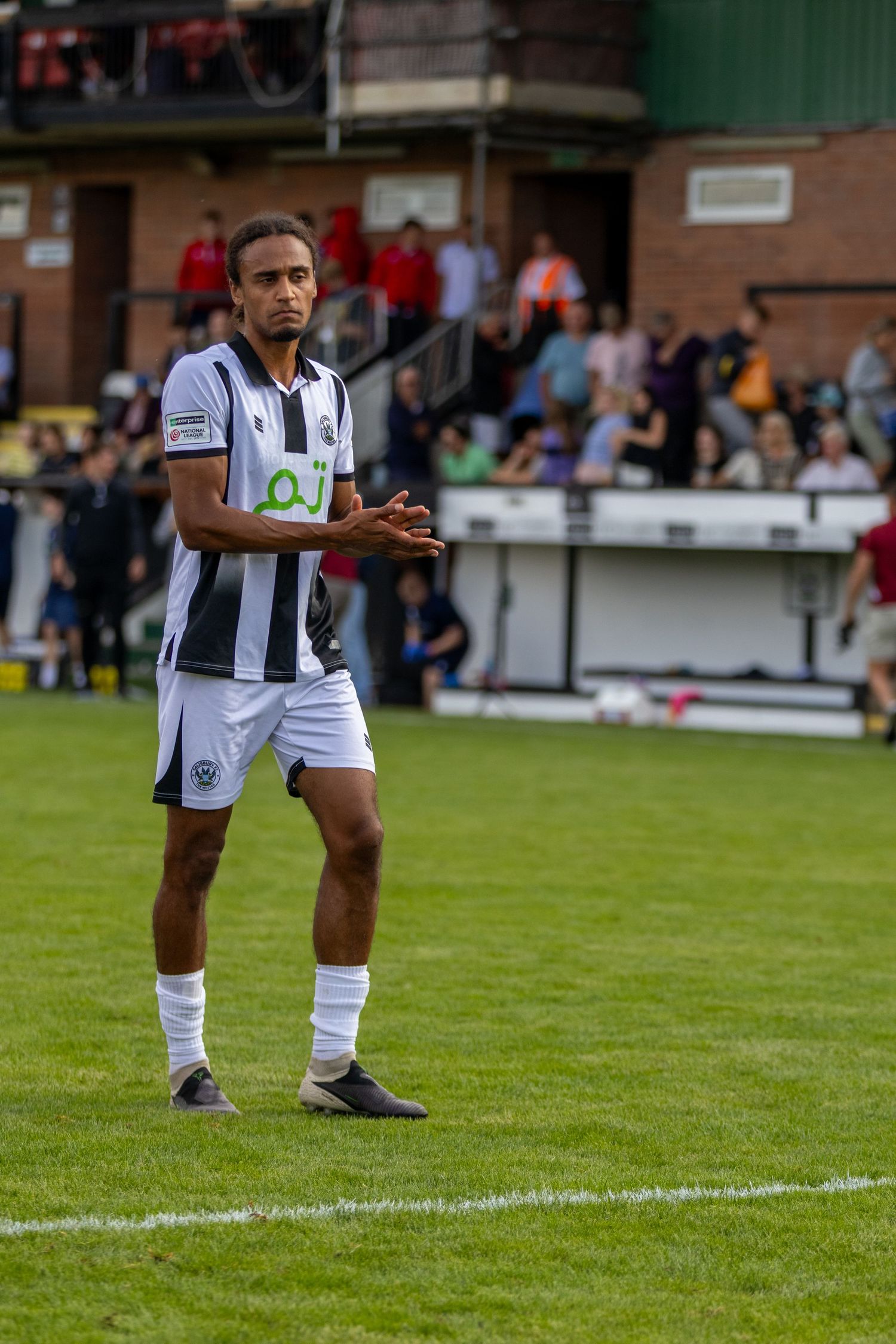 Soccer player in black and white striped kit clapping on a grass pitch during a match, with spectators in the background.