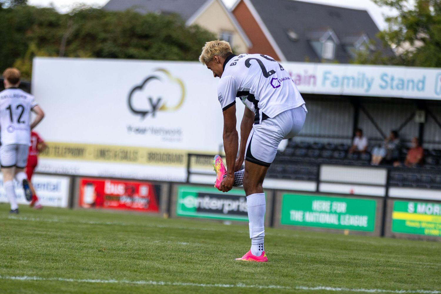 A player in white football kit stretches near the Retain Family Stand during a sunny outdoor match.