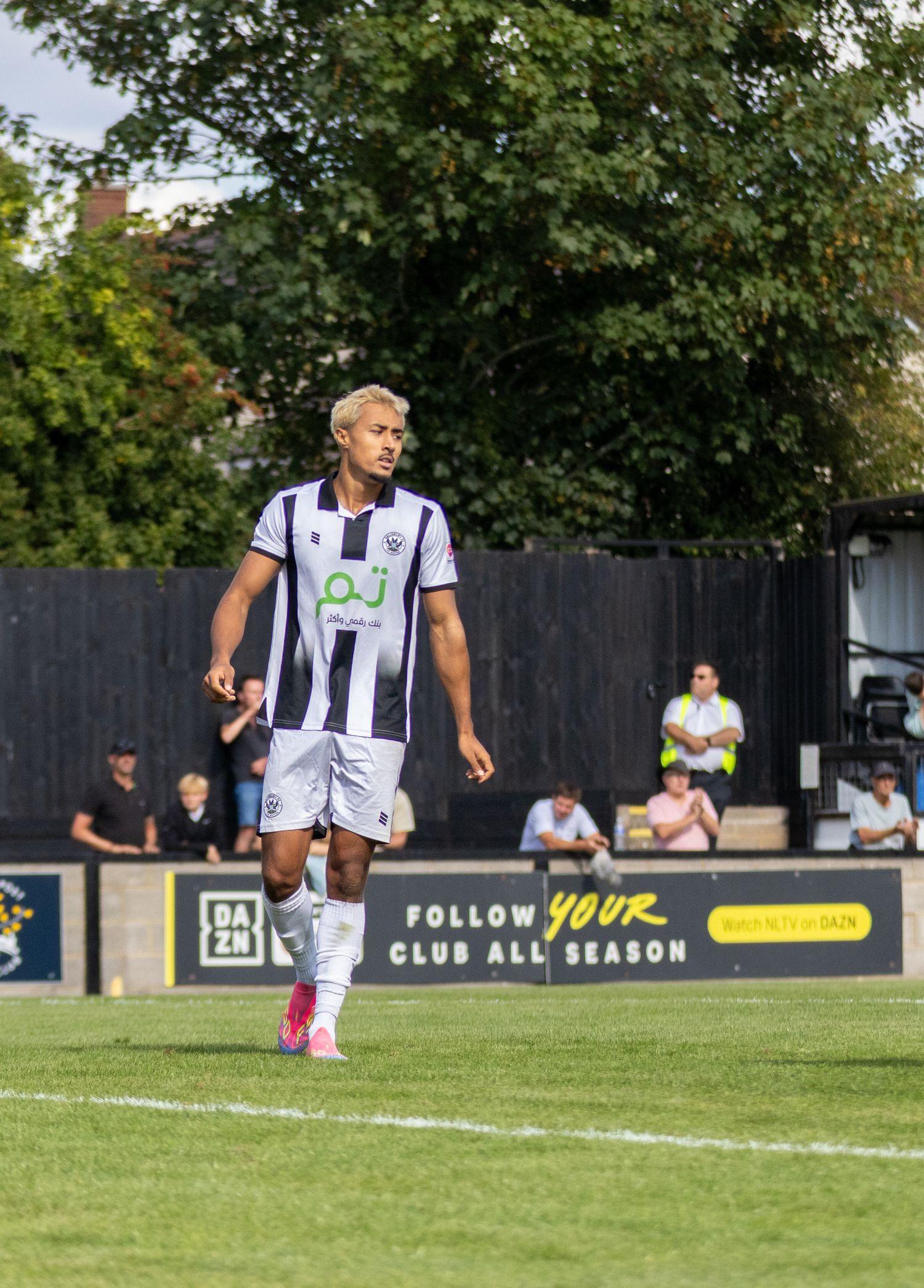 Blonde footballer in black and white striped kit walks on pitch near DAZN advertising board.