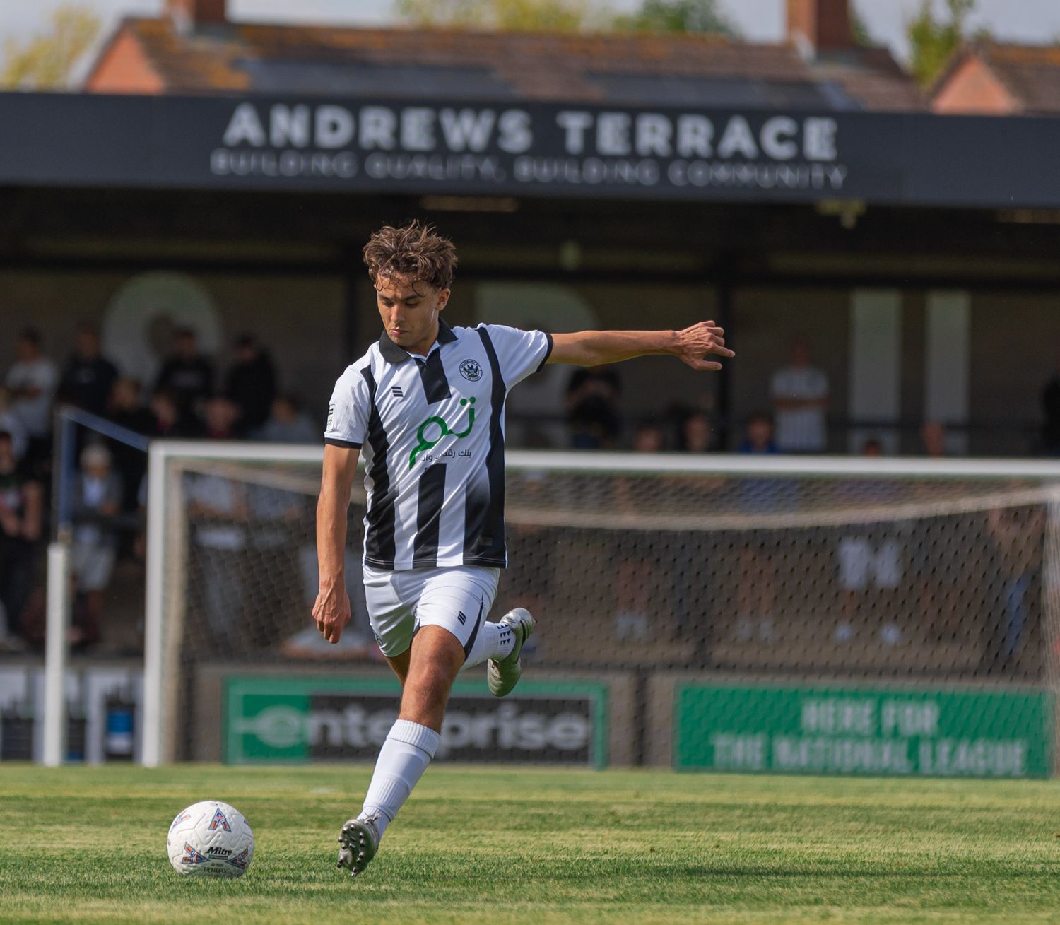 A footballer in black and white kit kicks a ball on a sunny pitch at Andrews Terrace ground.