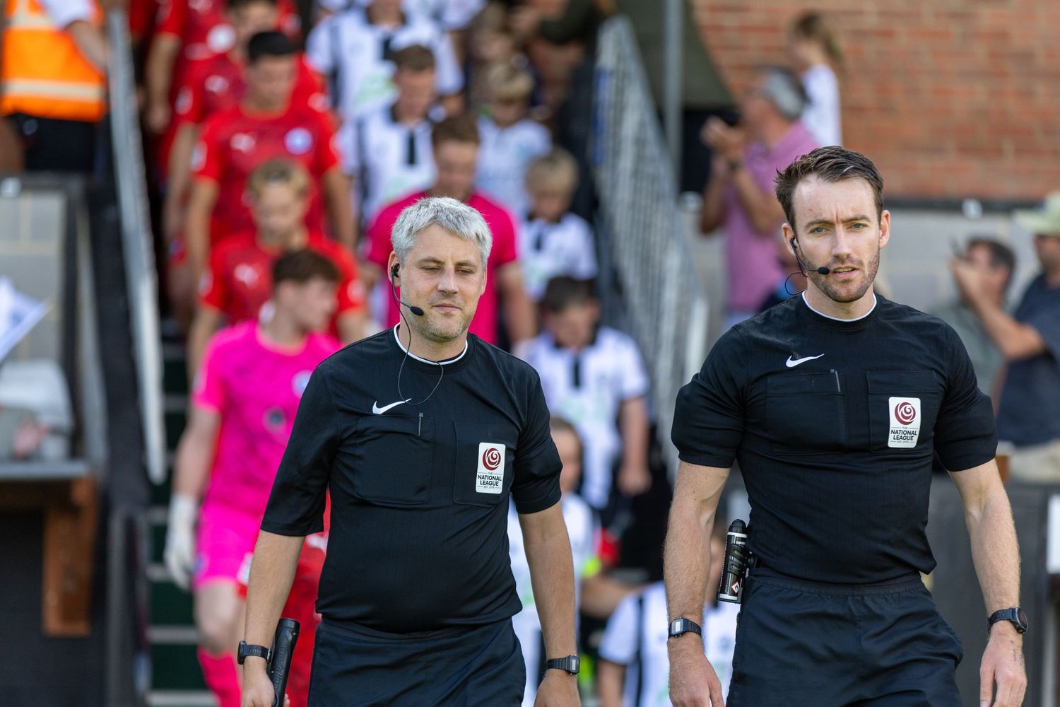 Two football referees in black uniforms walk onto the pitch before a match, with players visible in the background.