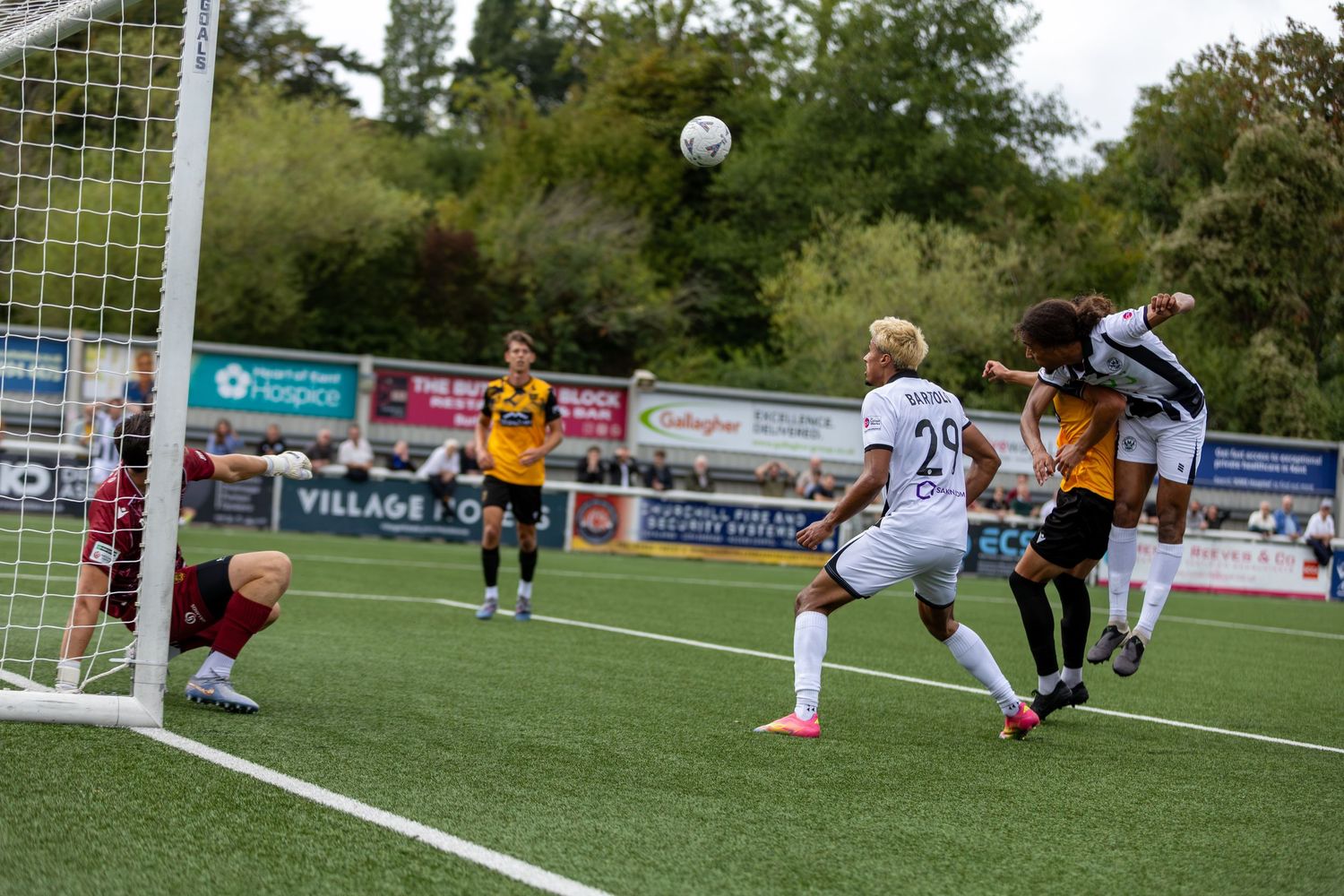 Soccer players compete for a header near the goal during an outdoor match, with spectators and advertising boards visible.