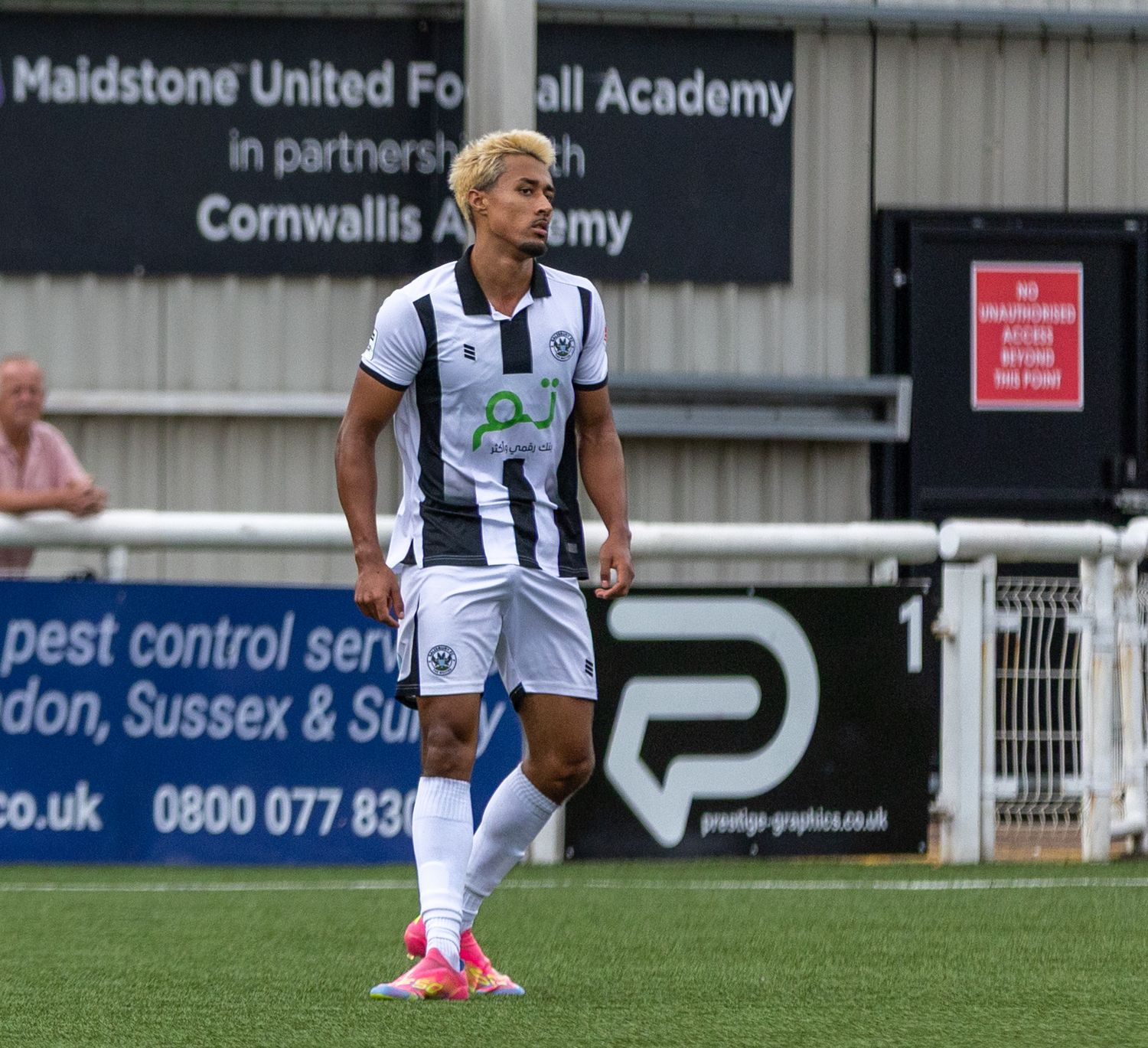 A footballer in black and white kit walks on the pitch at Maidstone United Football Academy during a match.
