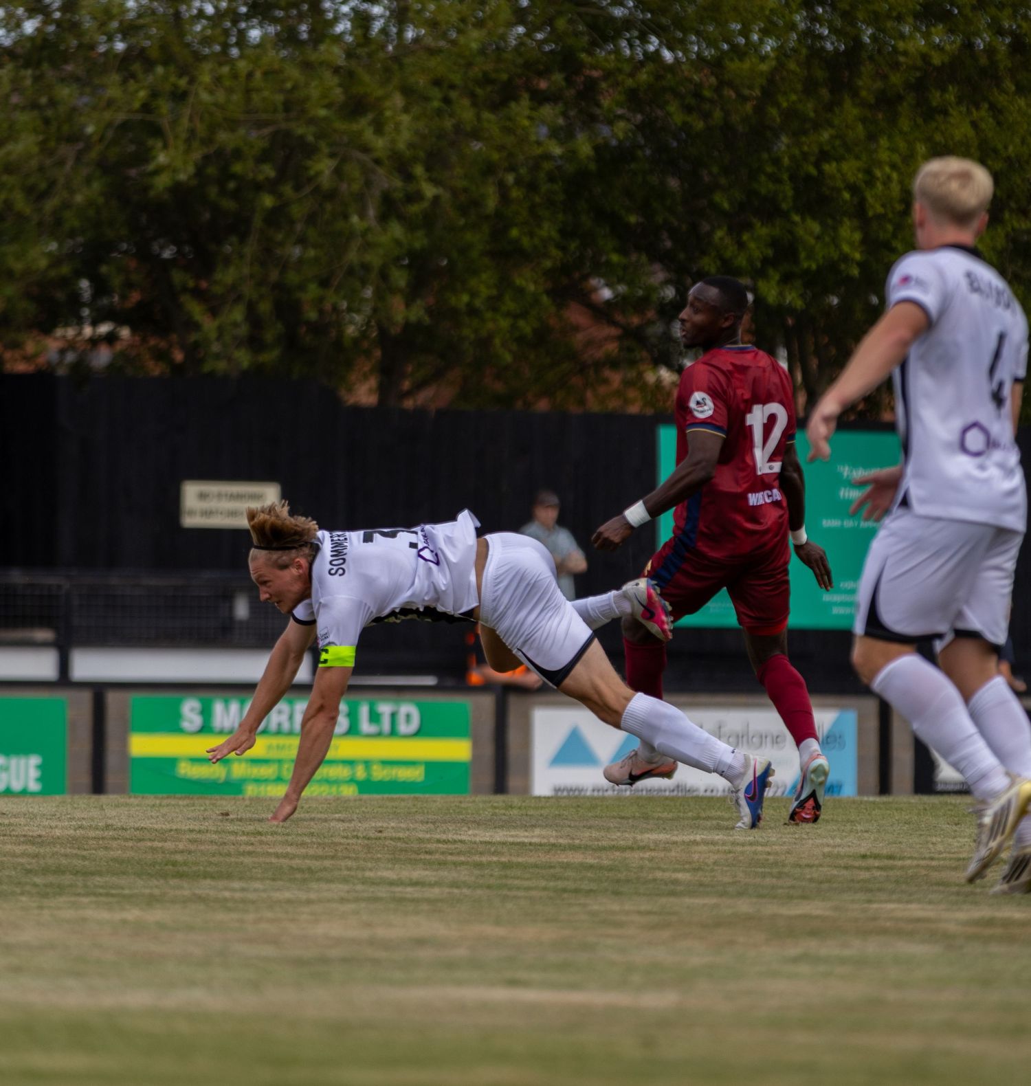 A footballer in white kit dives forward on a grass pitch as an opponent in red closes in during a match.
