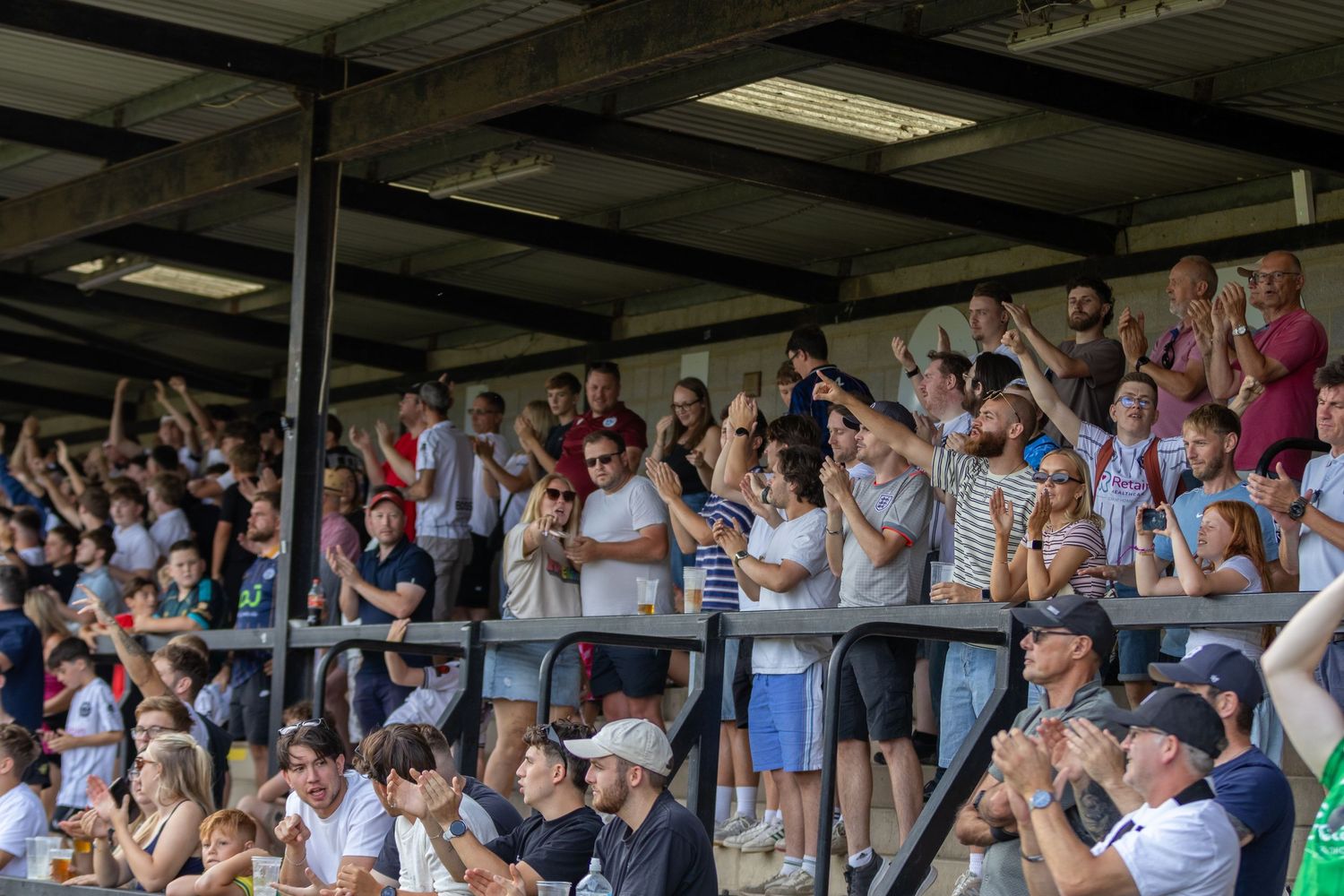 Large crowd of football fans cheering and clapping in a covered stadium stand on a sunny day.