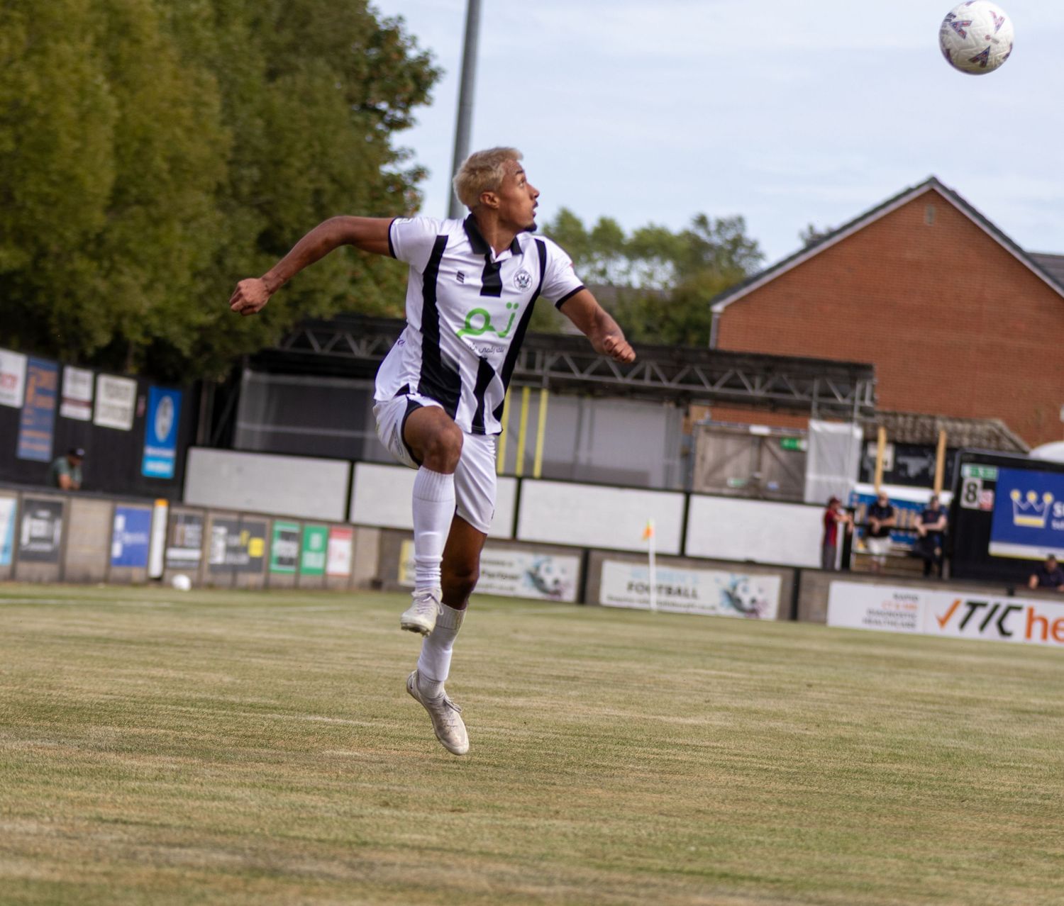 Soccer player in black and white kit leaps to head ball during sunny outdoor match near stadium stands.