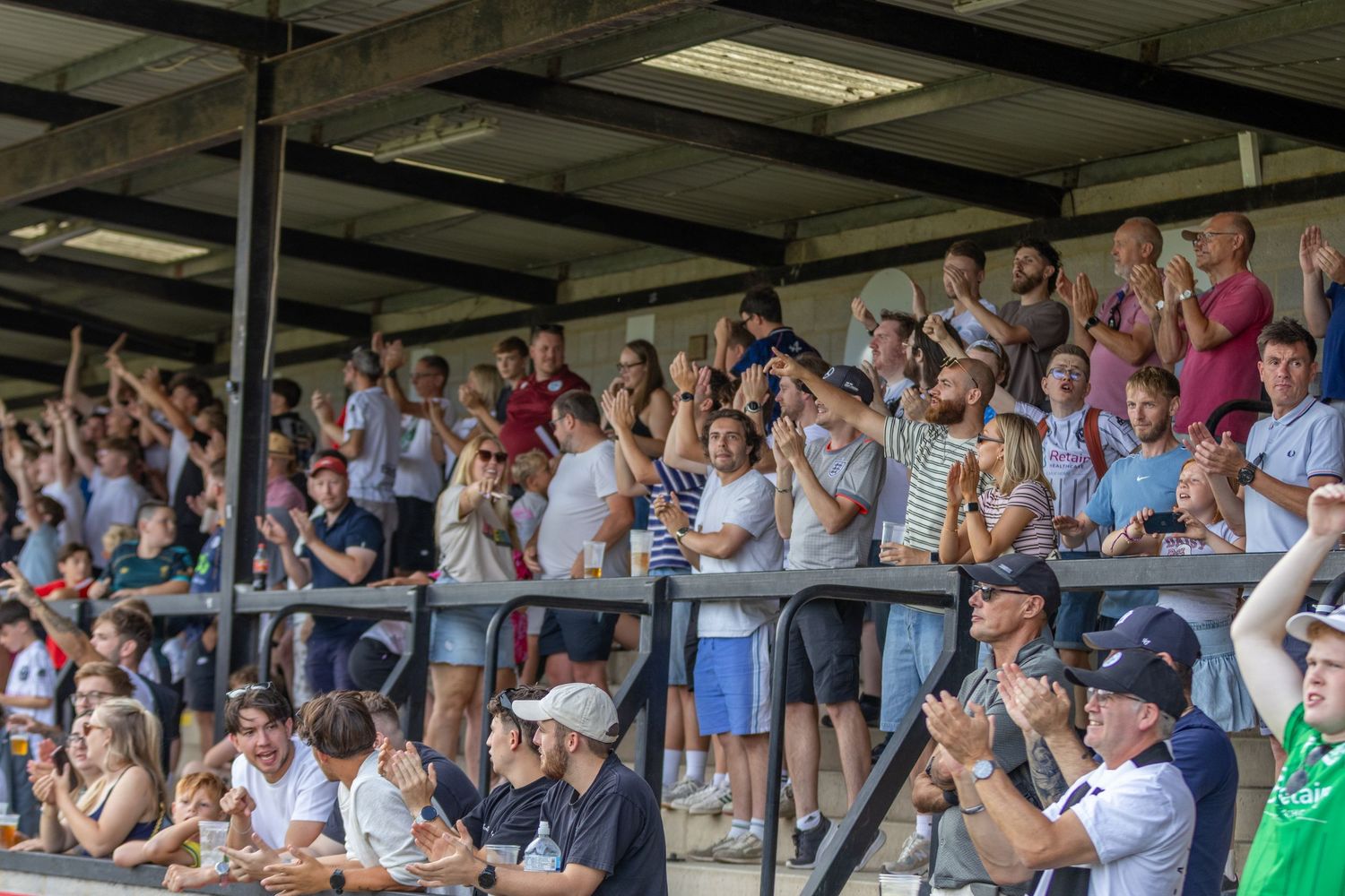 Excited football fans cheering in stadium stands on a sunny day at a non-league soccer match.
