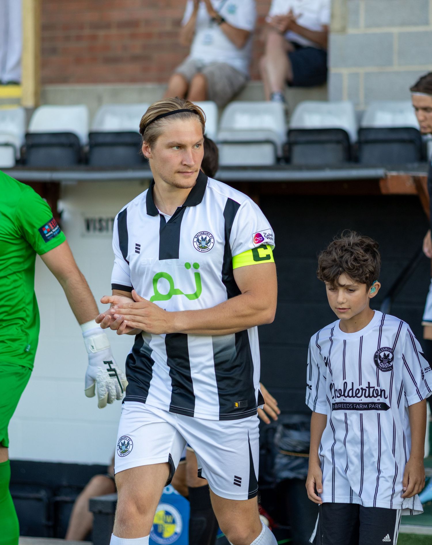 A footballer in a black and white striped kit shakes hands with a goalkeeper while a mascot walks beside him.