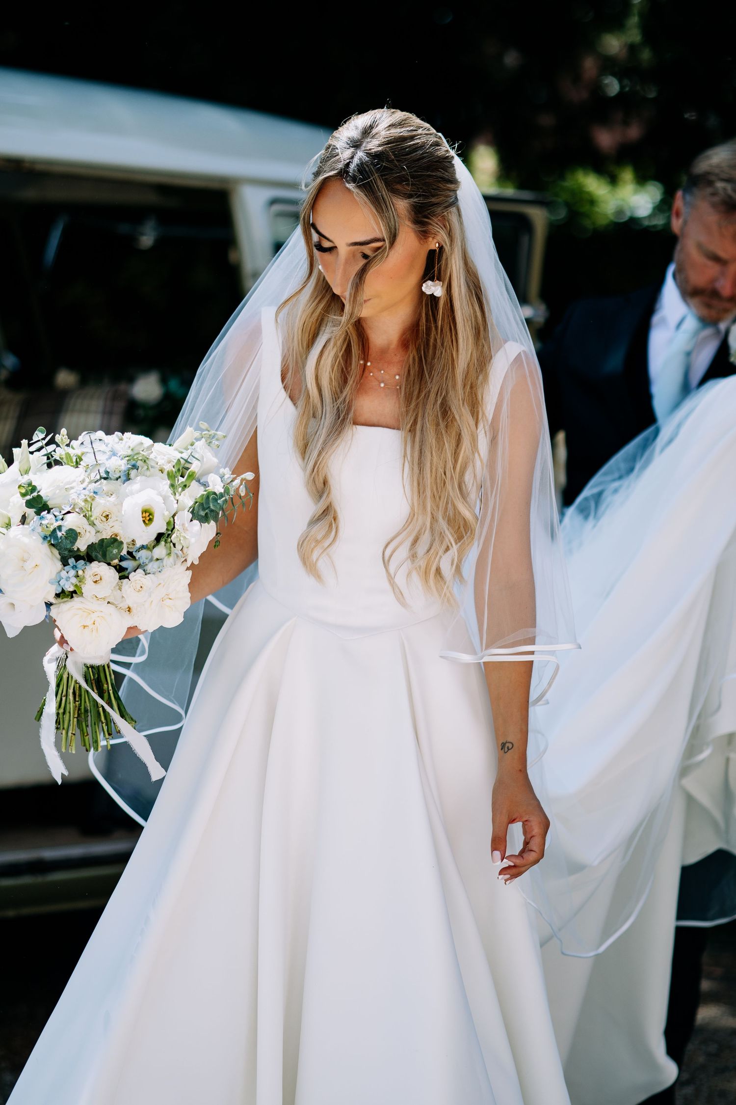 Elegant bride in white ball gown and veil holding white floral bouquet beside vintage car.