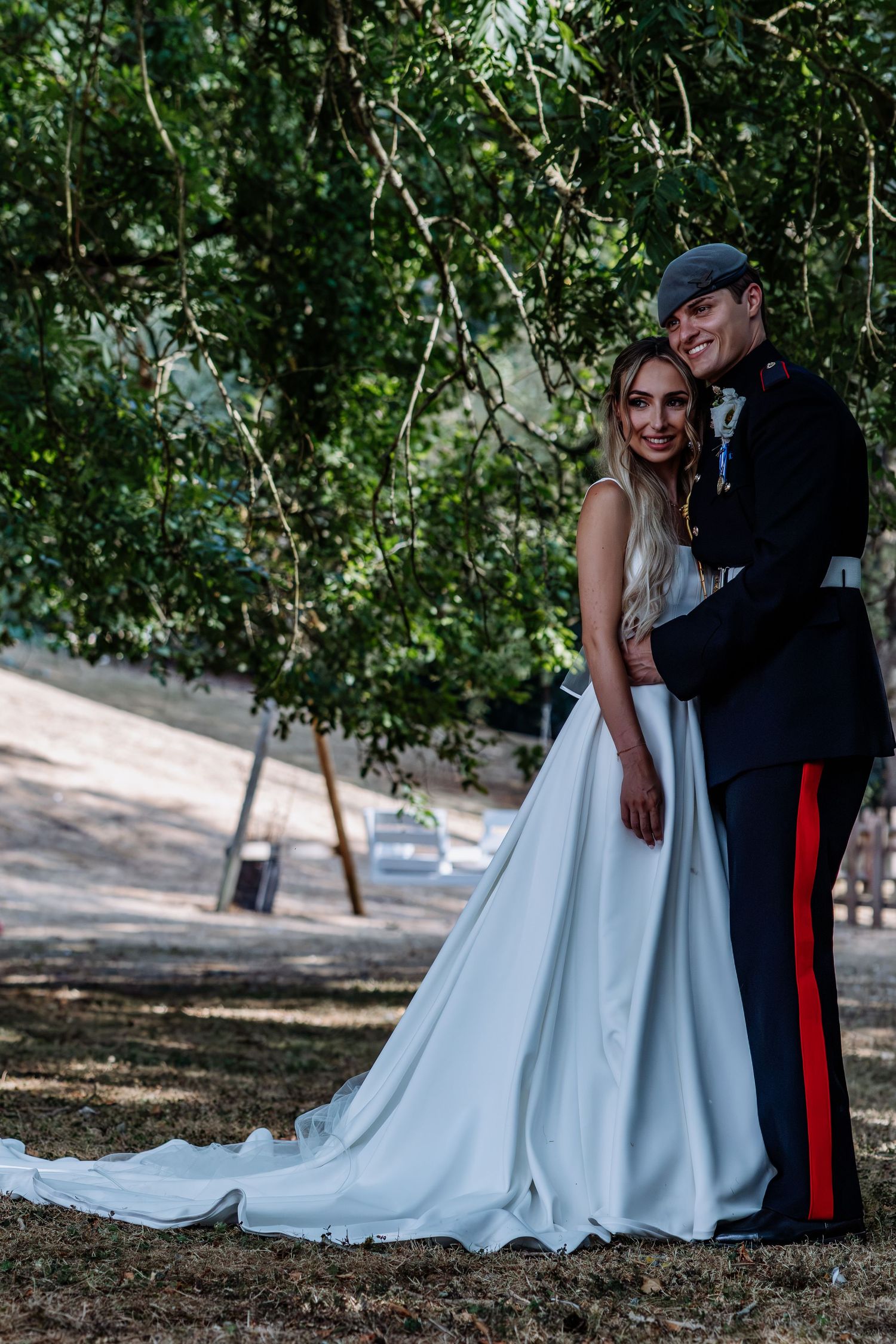 Bride in white gown and groom in military uniform pose under green trees.