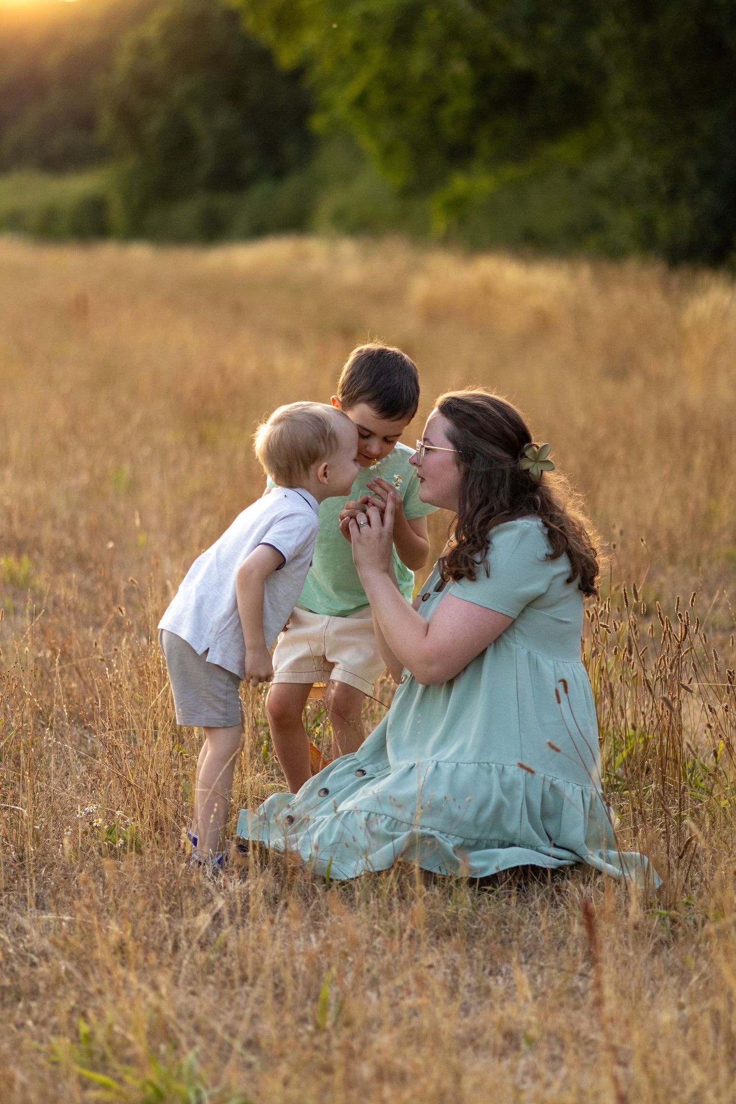 A pregnant woman in a teal dress sits in a golden field at sunset with two young boys leaning in for kisses.