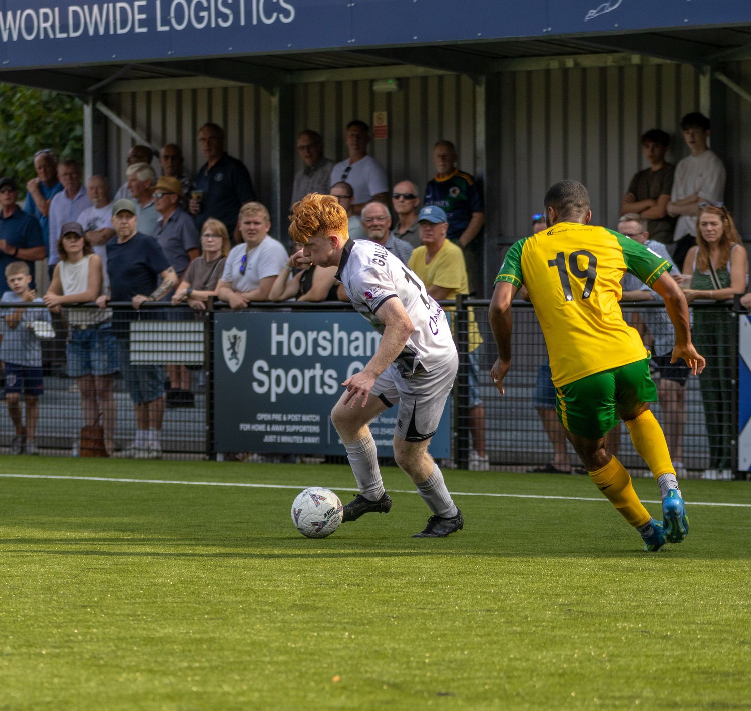 A red-haired footballer in white kit dribbles against a yellow-green opponent near the touchline during a match.