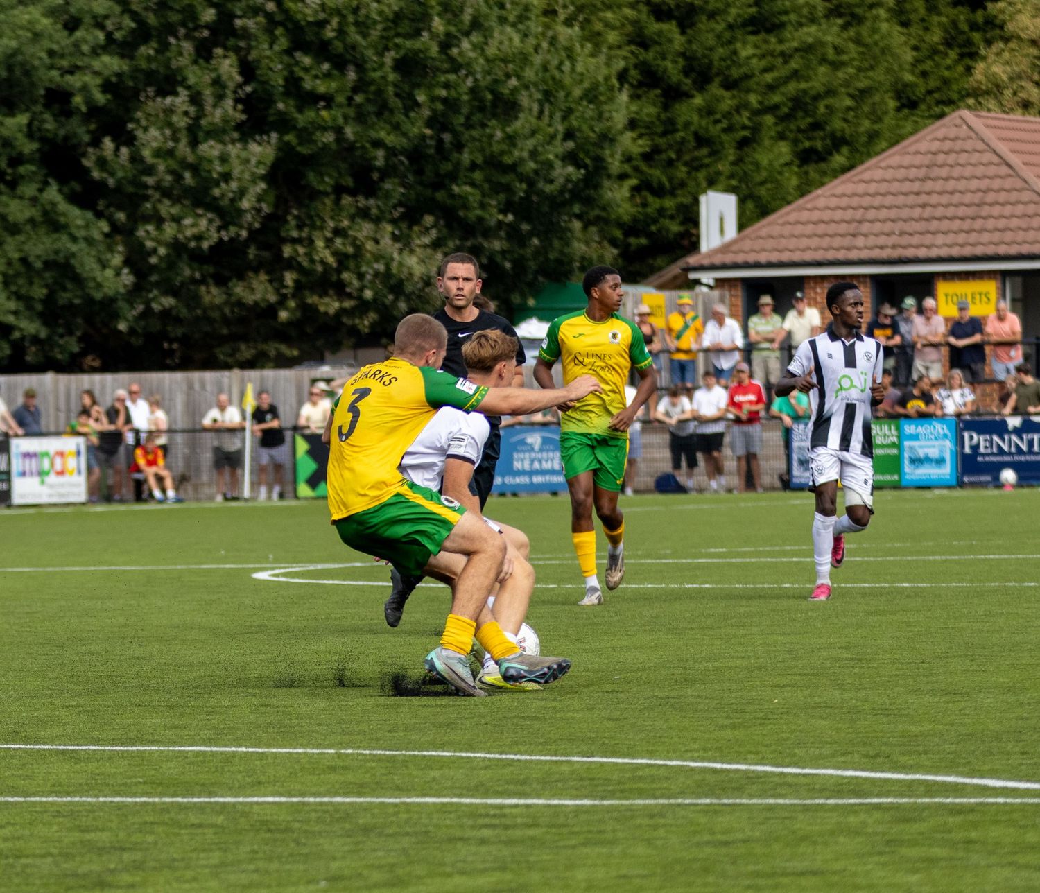 Two yellow-green footballers challenge a white-kitted player in a sliding tackle during a competitive non-league match.