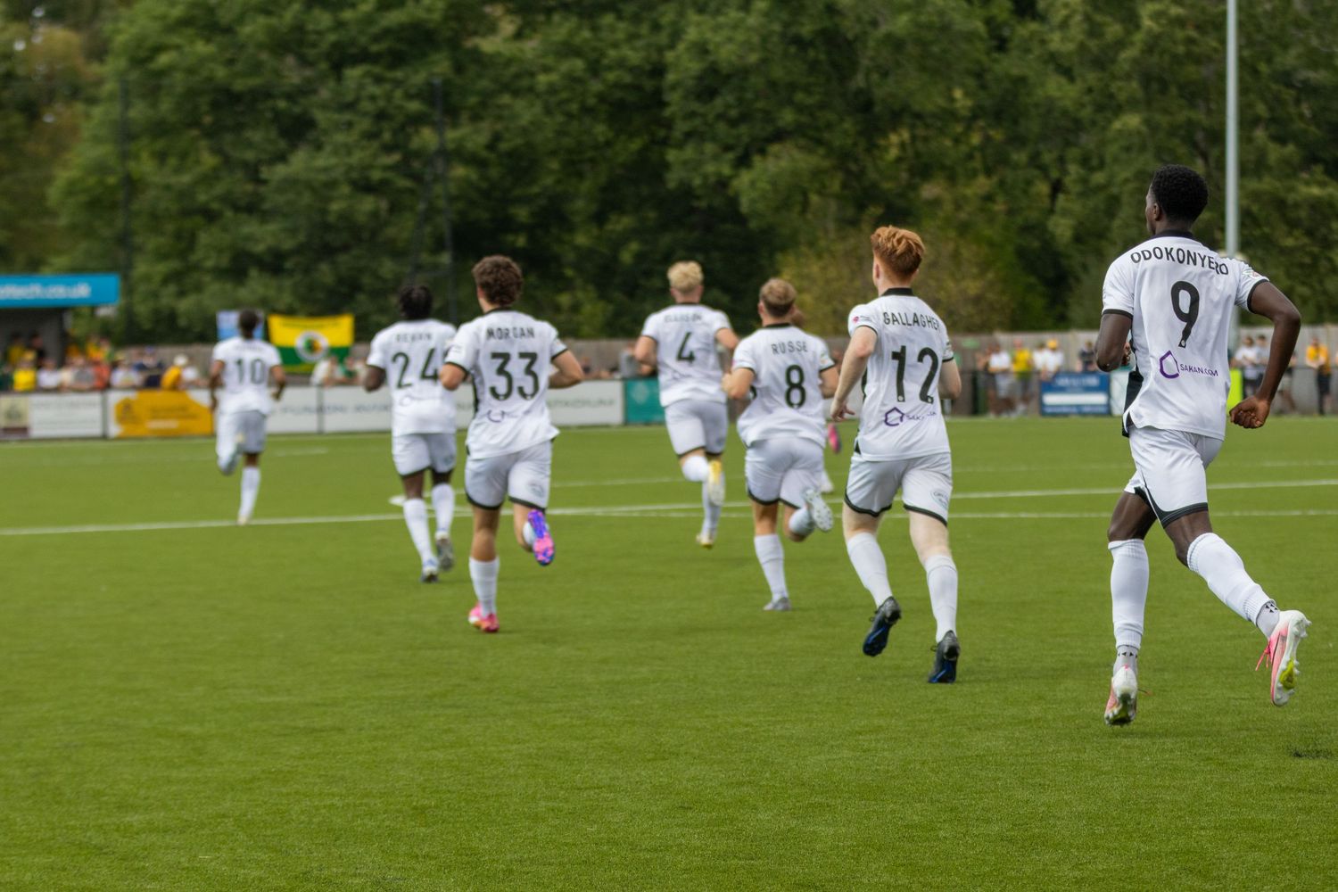 White-uniformed soccer players run across a green pitch during a competitive football match.