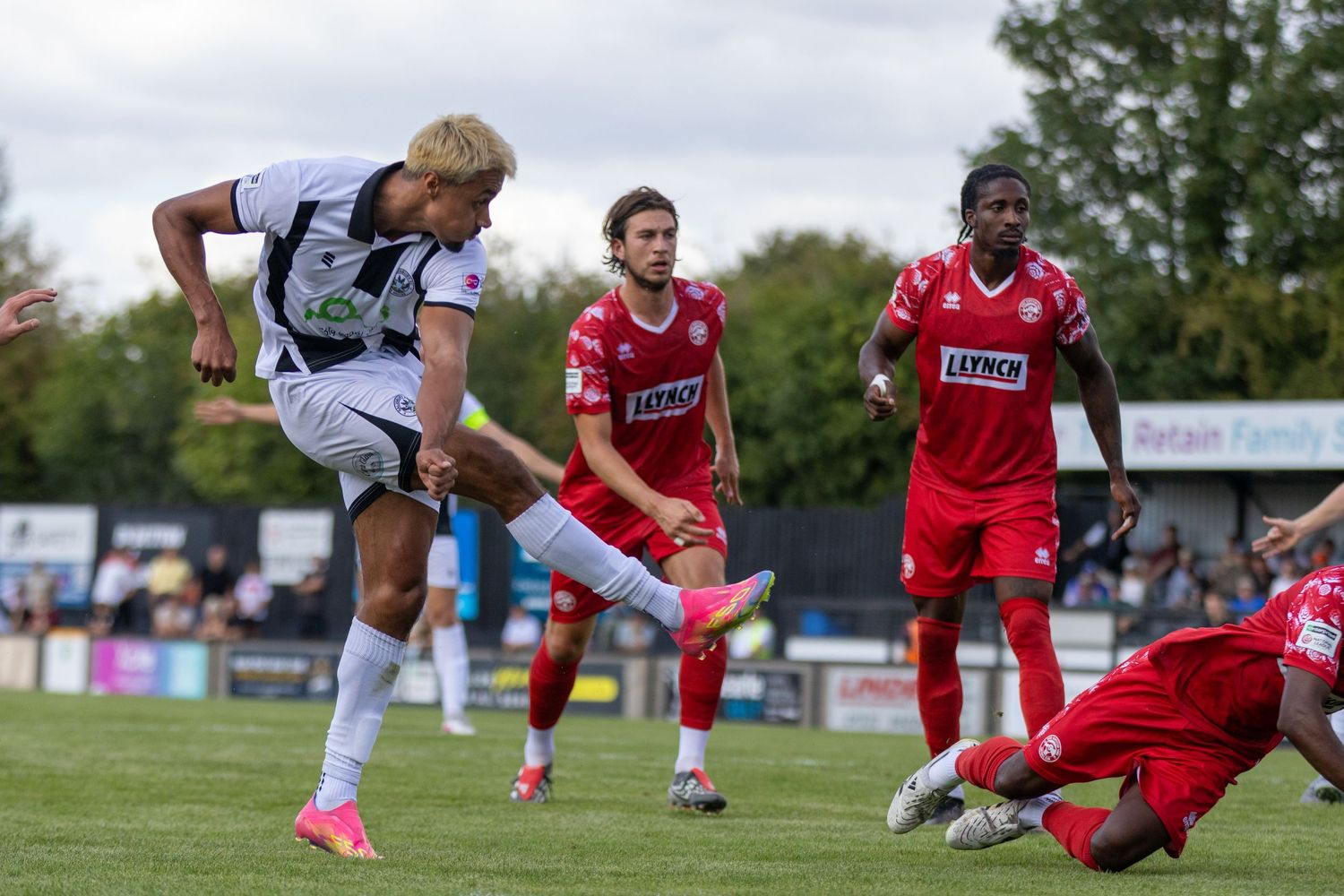 A player in black and white kit shoots at goal during a match against red-kitted opponents.