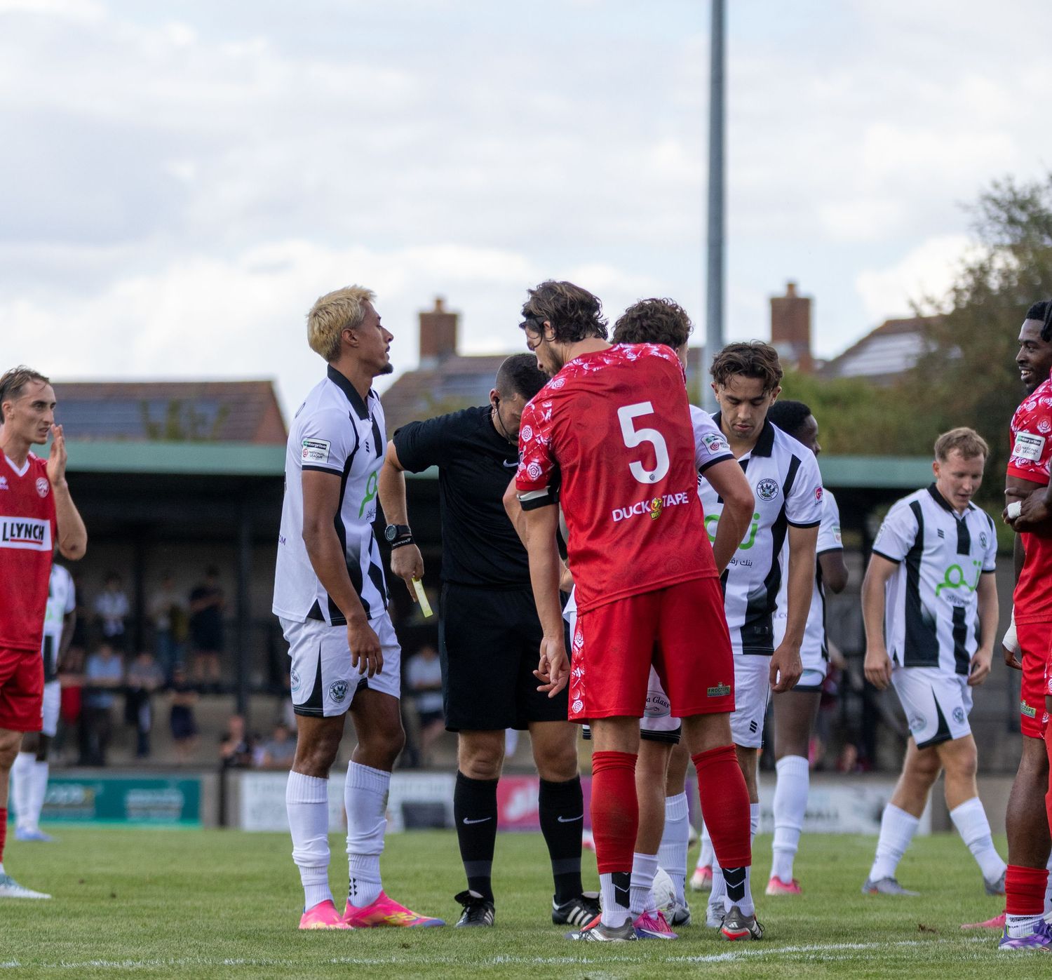 Referee speaks to players in red and white kits during a non-league football match on a cloudy day.