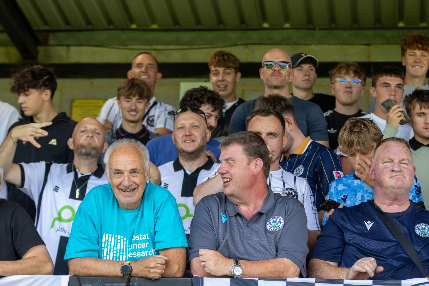 Football supporters laughing and cheering together in packed stadium stands on a bright sunny day.