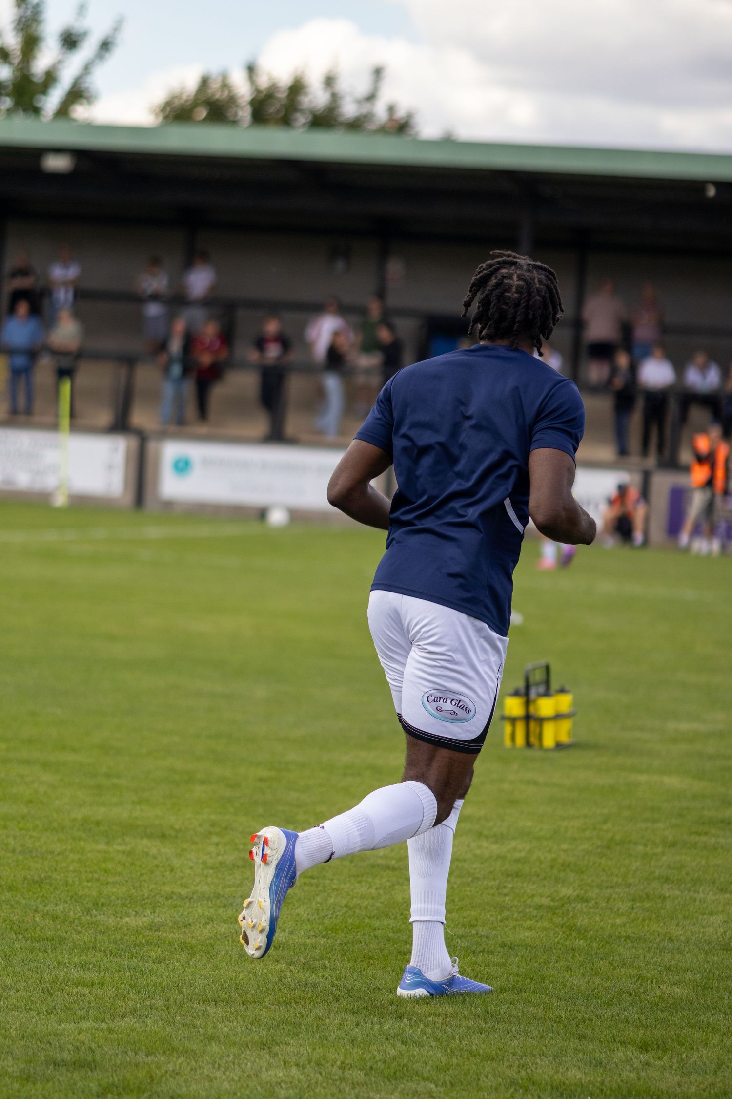 A footballer in navy blue shirt and white shorts runs away from camera during warm-up on a green football pitch.
