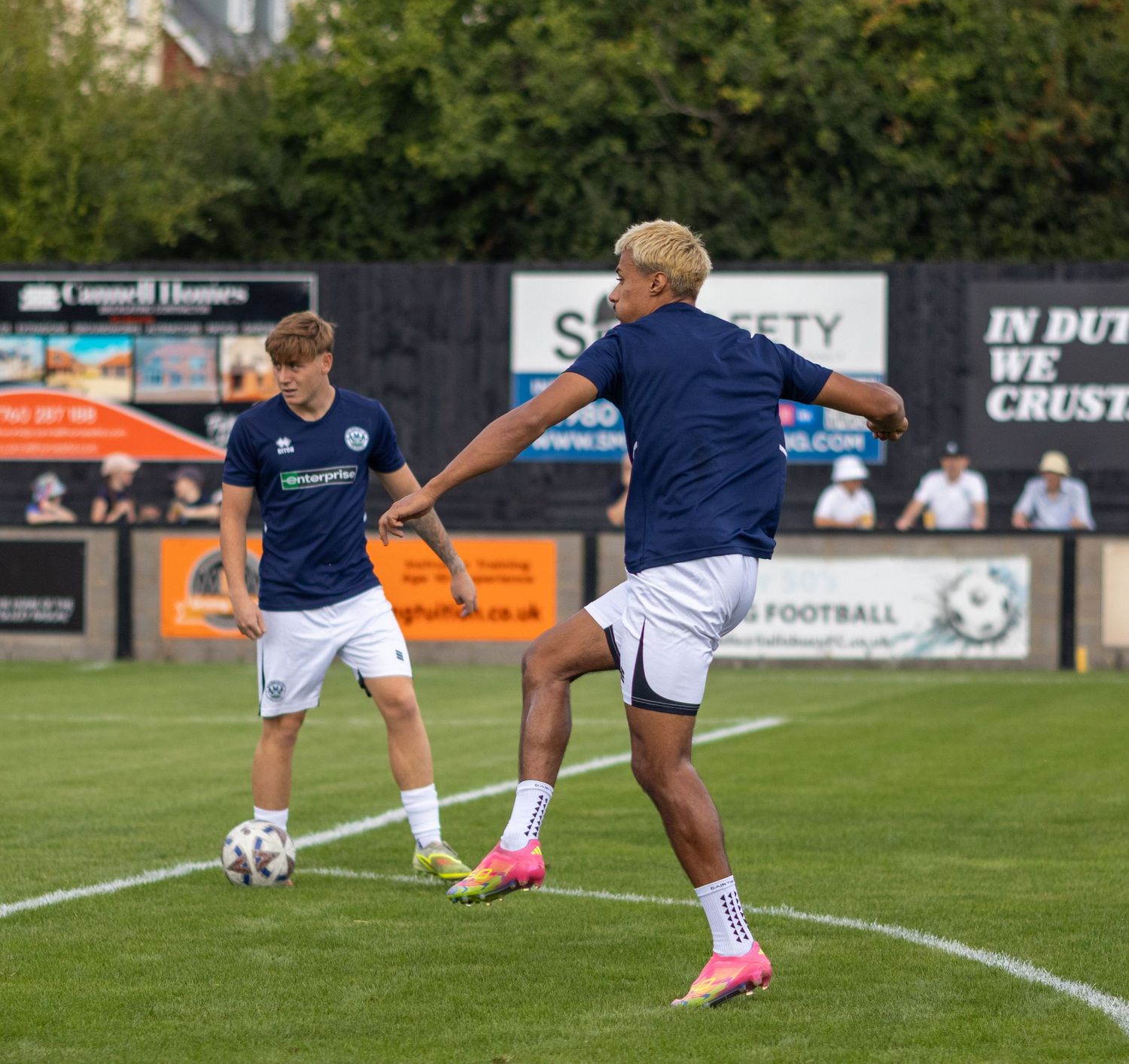 Two football players in navy blue kits warm up on a grass pitch during a pre-match training session.
