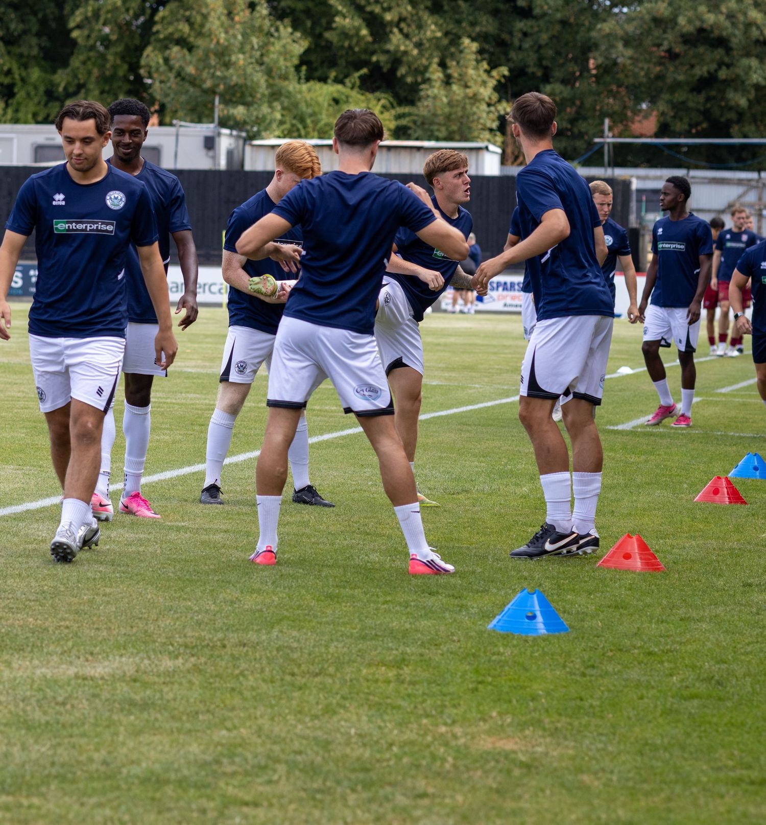 Football players in navy Enterprise-sponsored kits warm up with cones on a grass pitch before a match.