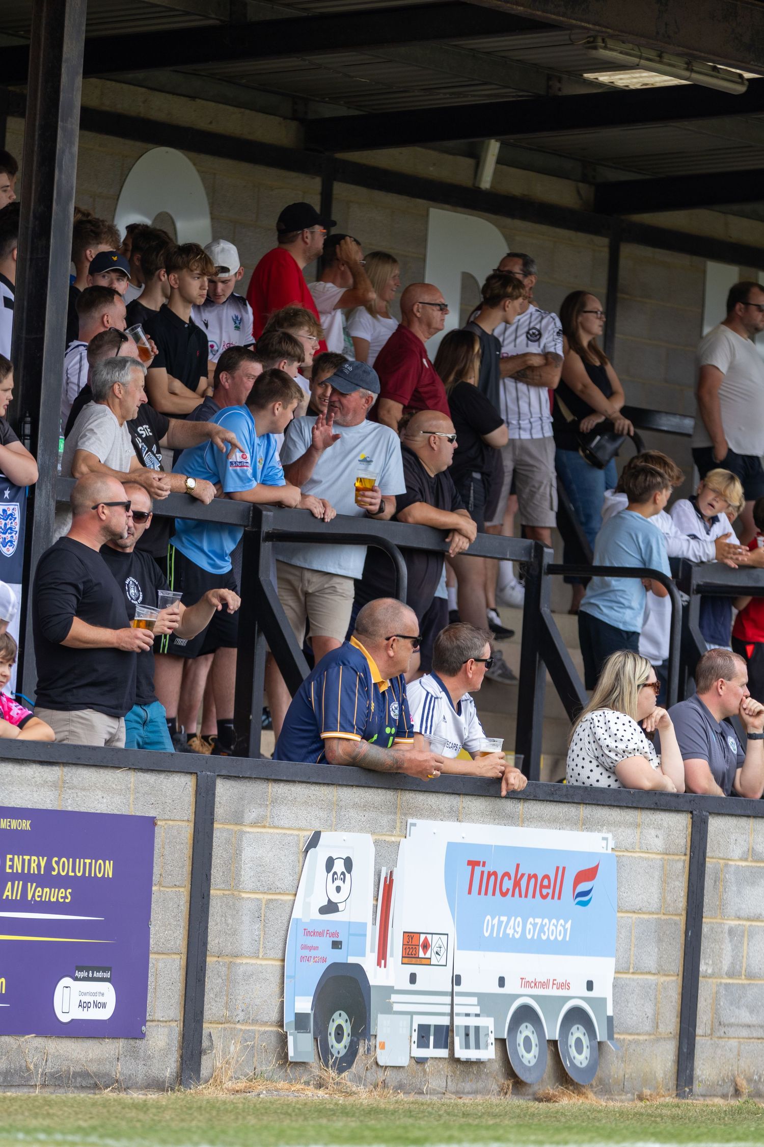 Football fans gather in a covered stand drinking and watching a non-league match with sponsor signage visible.