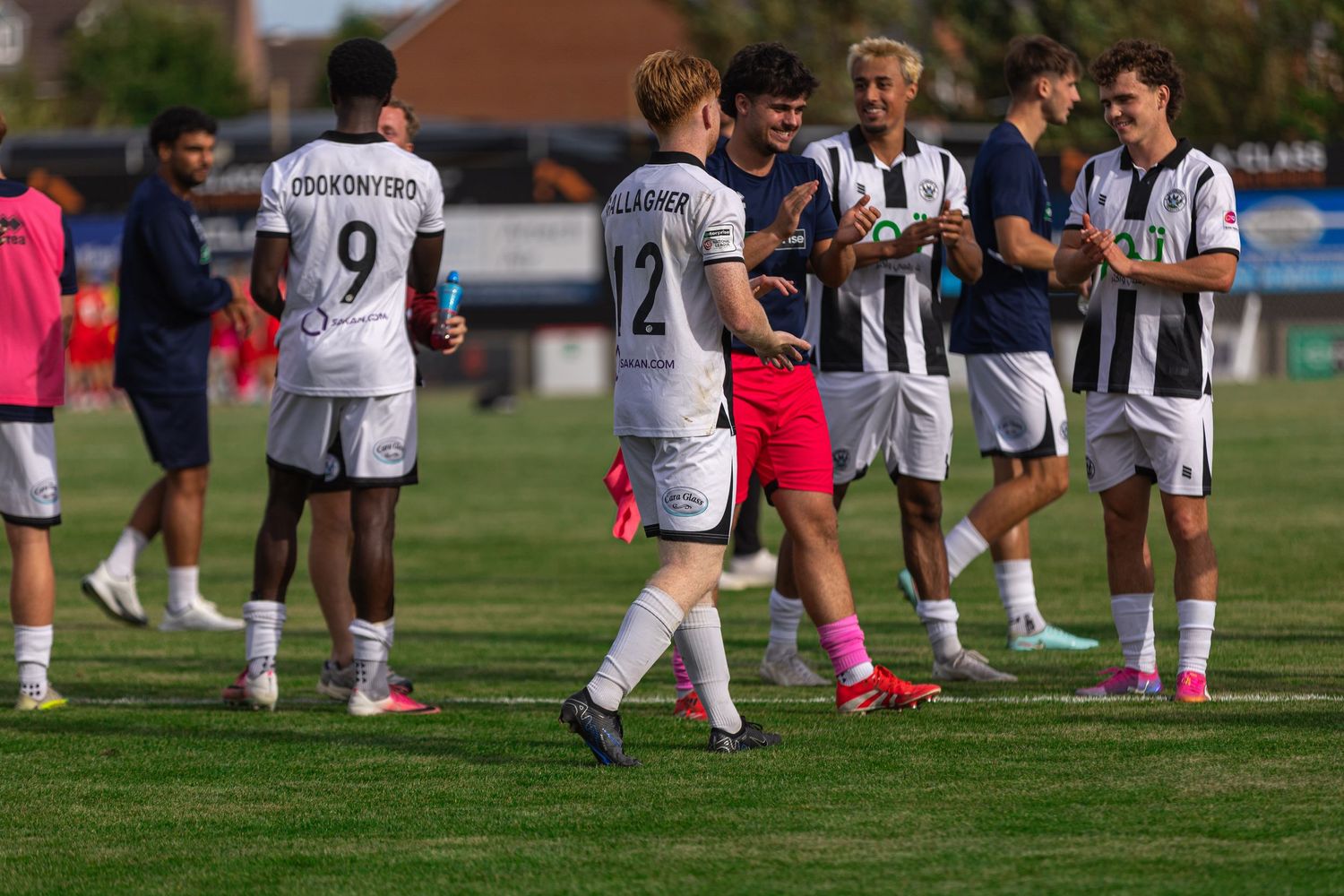Soccer players in black and white uniforms celebrate on a green field during a sunny outdoor match.