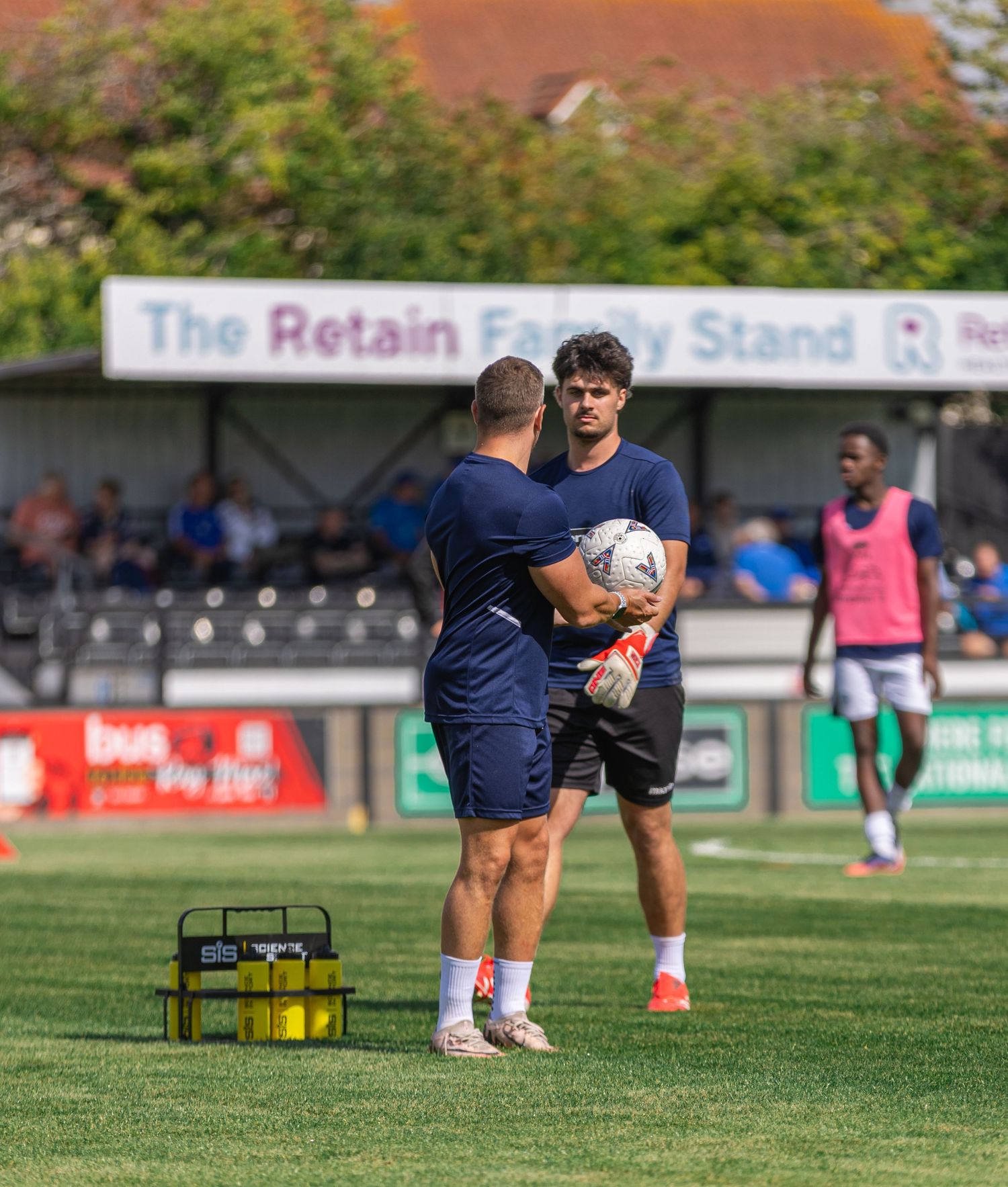 Two footballers training on a pitch in front of The Retain Family Stand on a sunny day.