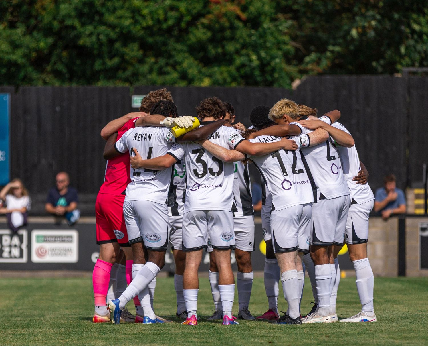 Soccer team huddles together on green field before match, wearing white uniforms on a sunny day.