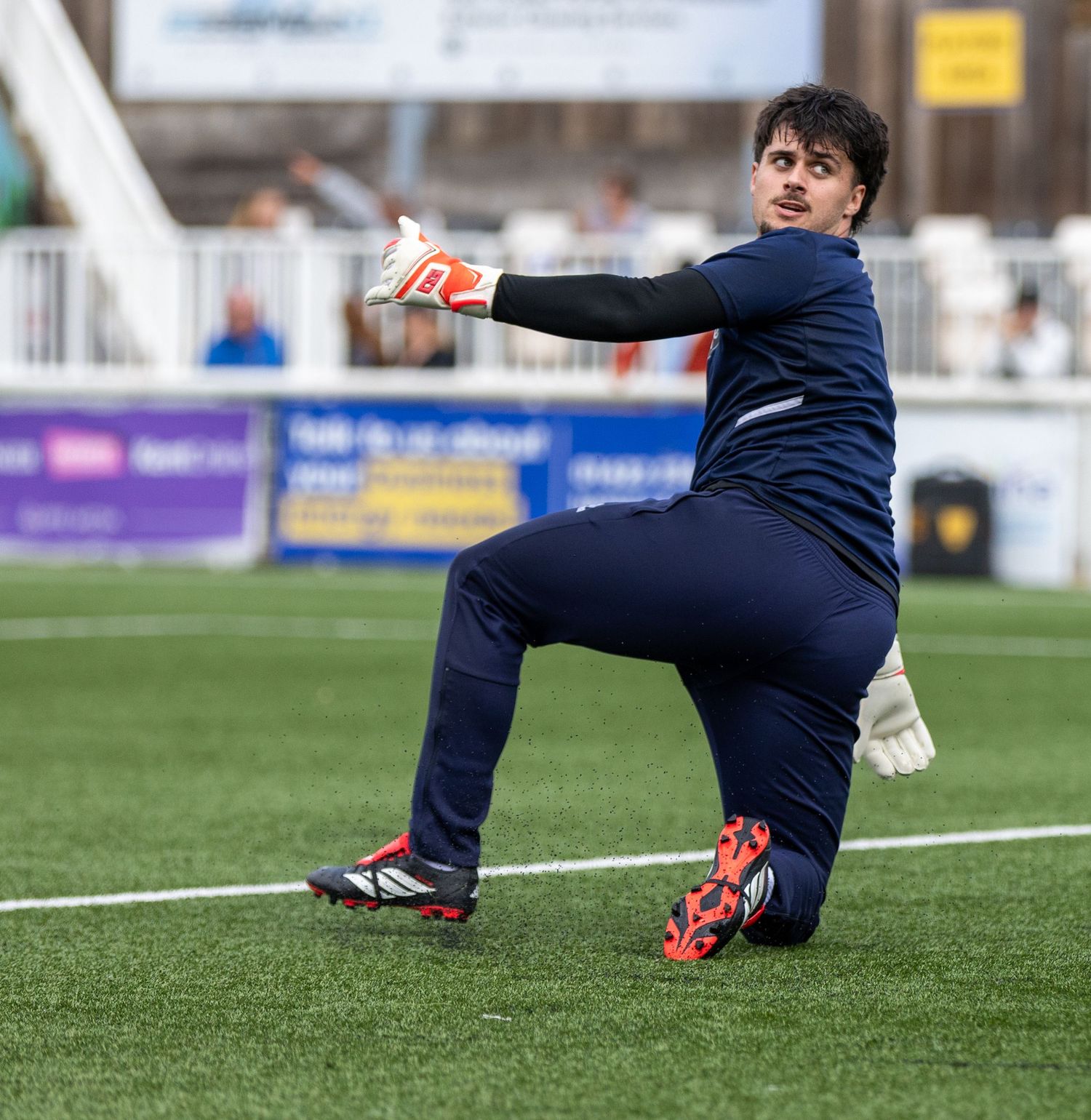 A goalkeeper in navy blue kit and orange gloves stretches during warm-up on a green football pitch.