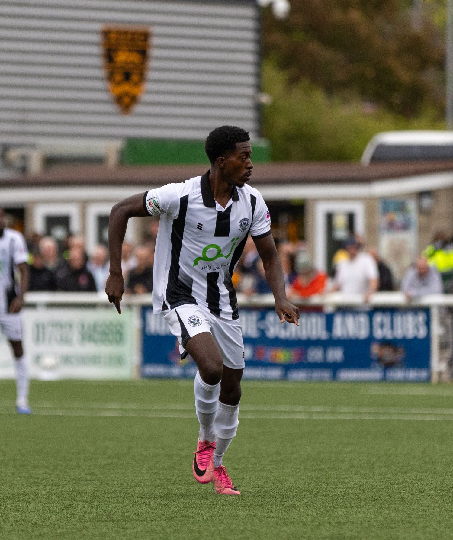 A young male soccer player in black and white striped kit with pink cleats running on a grass field during a match.