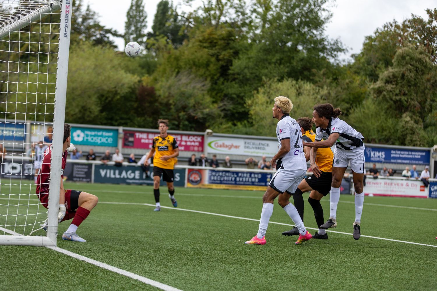 Soccer players compete near the goal during an outdoor match on a green pitch with trees in the background.