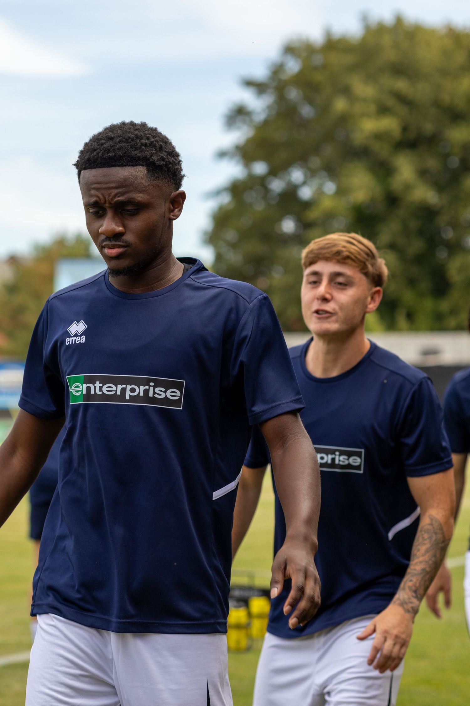 Two Amesbury FC players in navy Enterprise-sponsored Errea kits walk on the pitch during pre-match warmup.