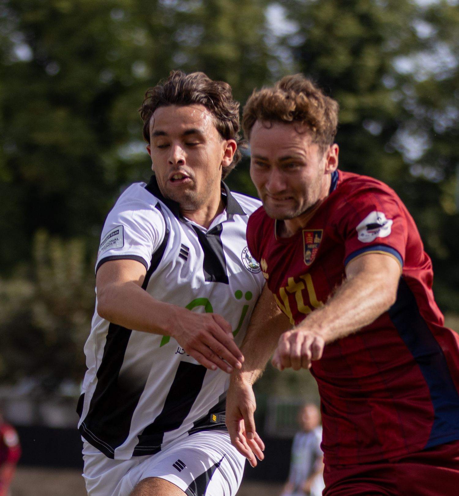 Two footballers in black-white and red kits challenge for the ball in a close non-league match duel.