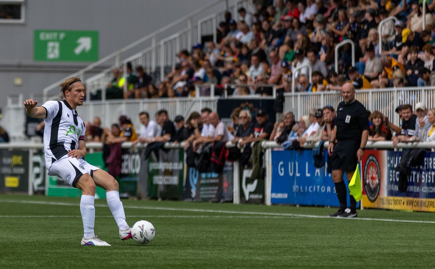 A footballer in white kit prepares to kick the ball during a match, with a referee and packed stands visible in the background.