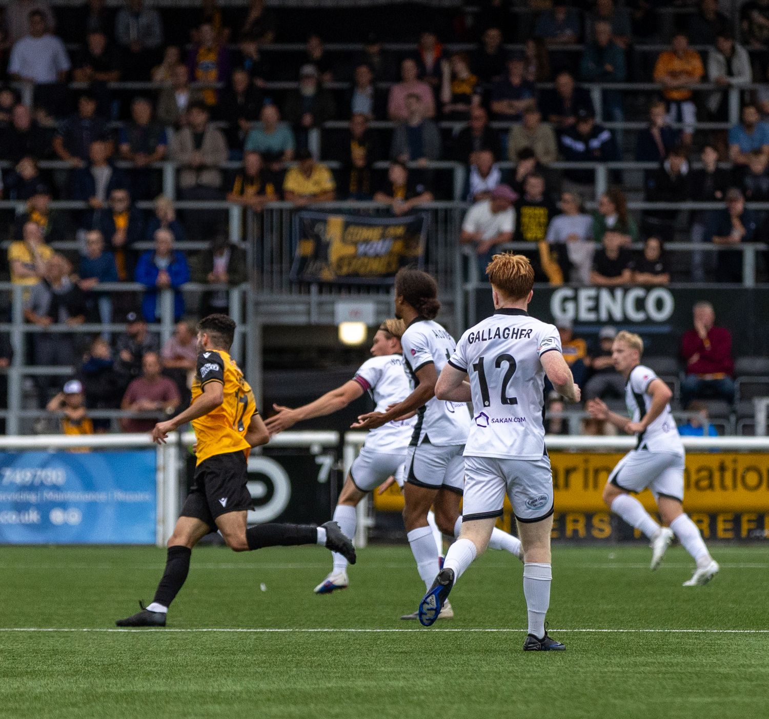 Football players in white jerseys celebrate during a match, with fans in stadium stands visible in the background.