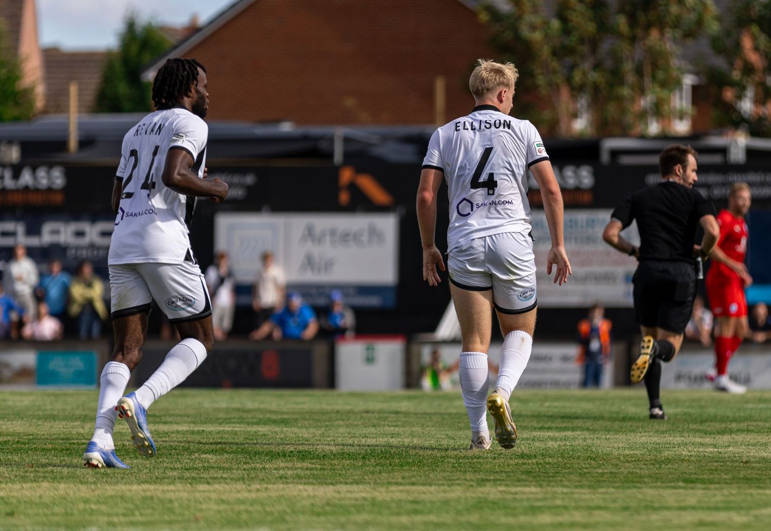 Two soccer players in white uniforms, numbers 21 and 4, celebrate on a sunny outdoor pitch during a match.